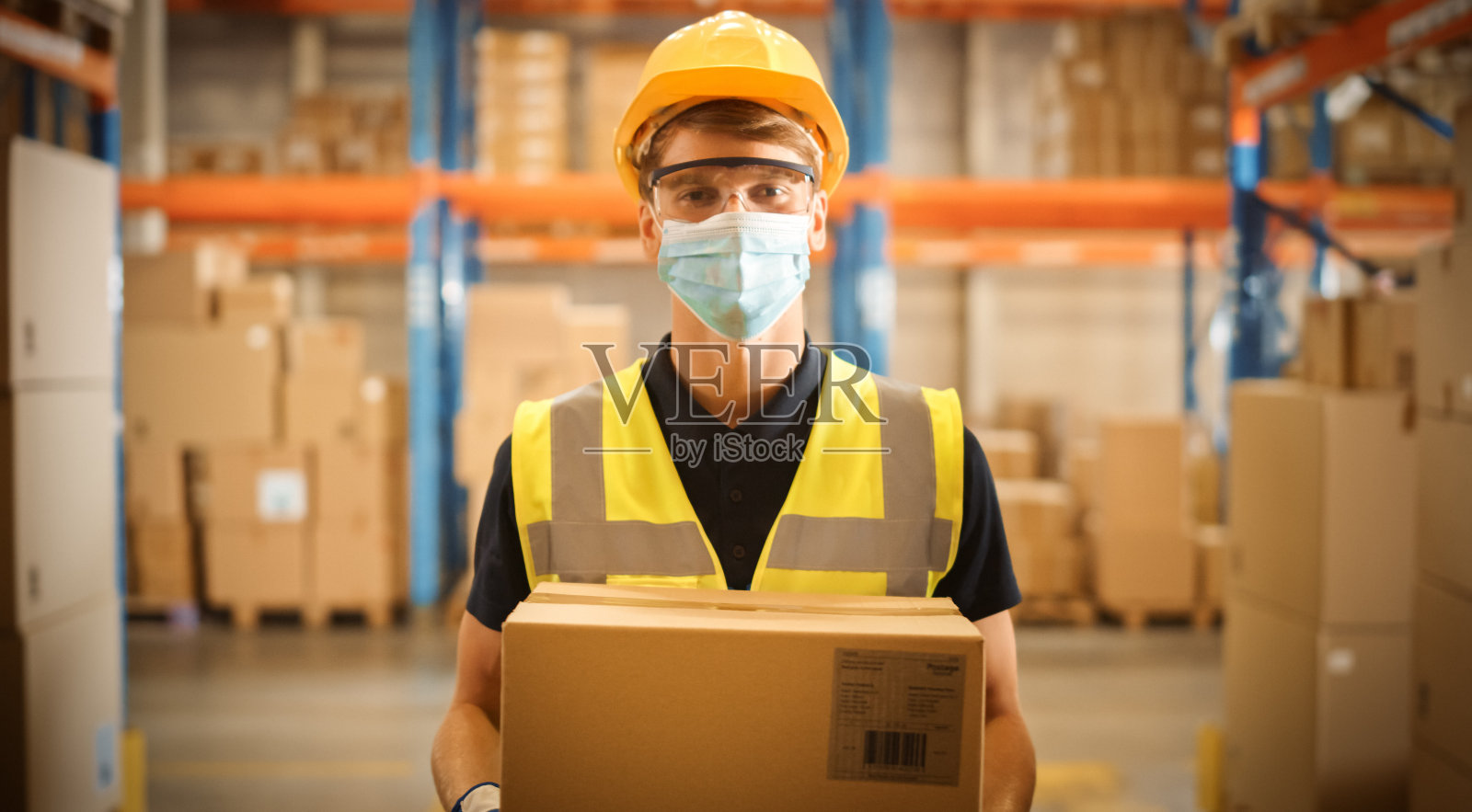 帅气的男工人戴着医用口罩和安全帽，手持纸板箱，站在摆满货架的大型零售仓库里。安全第一防护工作场所照片摄影图片
