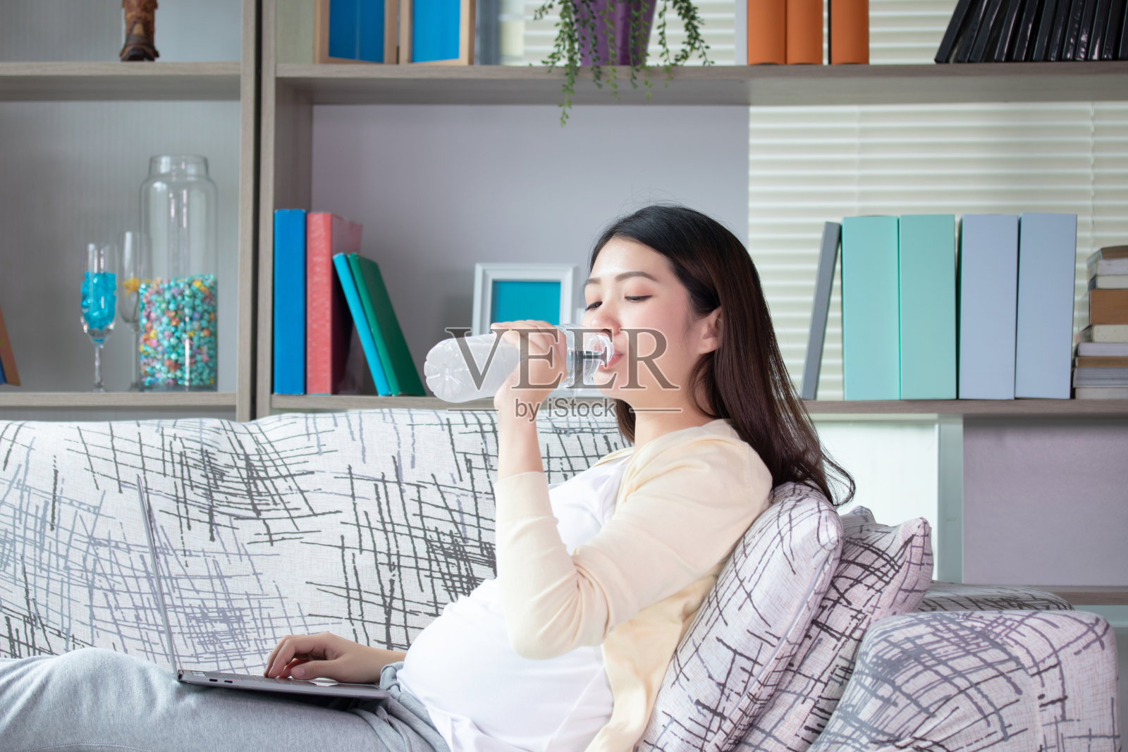 年轻漂亮的孕妇坐在客厅的沙发上喝着塑料瓶里的水。放松、休息的时间观念。照片摄影图片