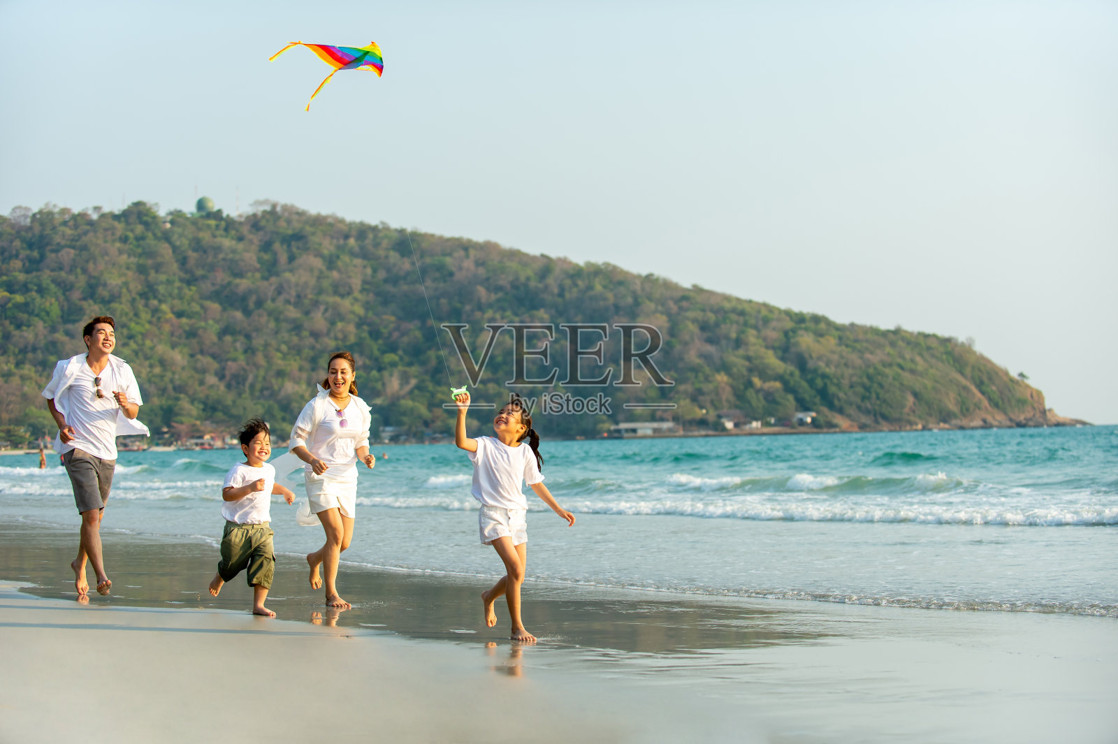 快乐的亚洲家庭父母和两个孩子跑步和玩风筝一起在夏天的时间在海滩上日落。父亲，母亲和孩子们放松和享受夏季户外生活方式假期。照片摄影图片