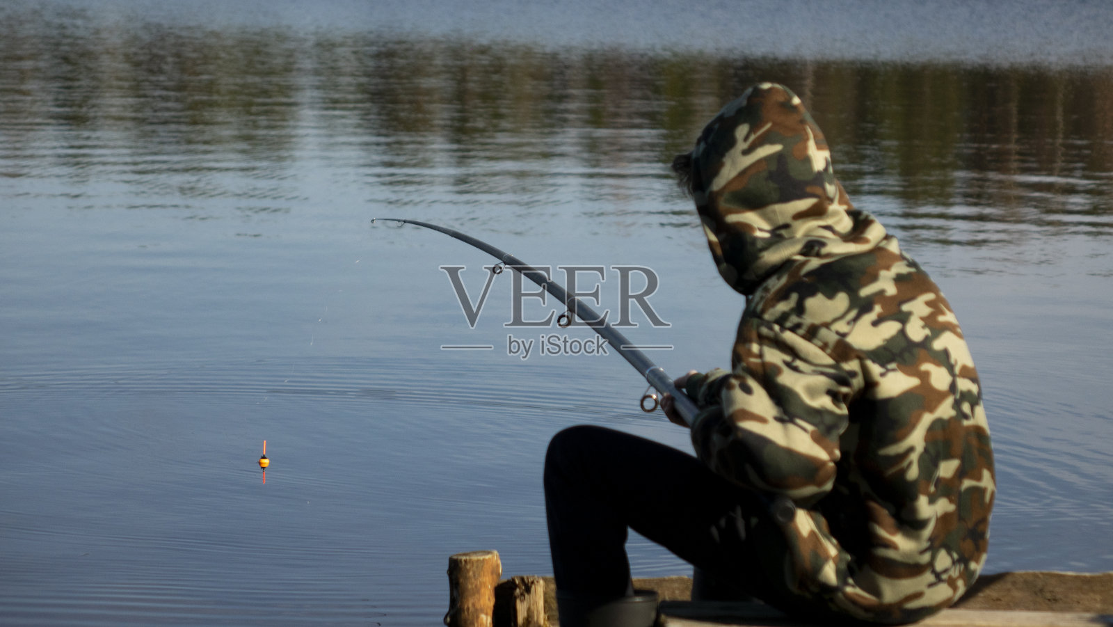 湖岸边穿着迷彩服的渔夫。用浮杆在水上钓鱼。照片摄影图片