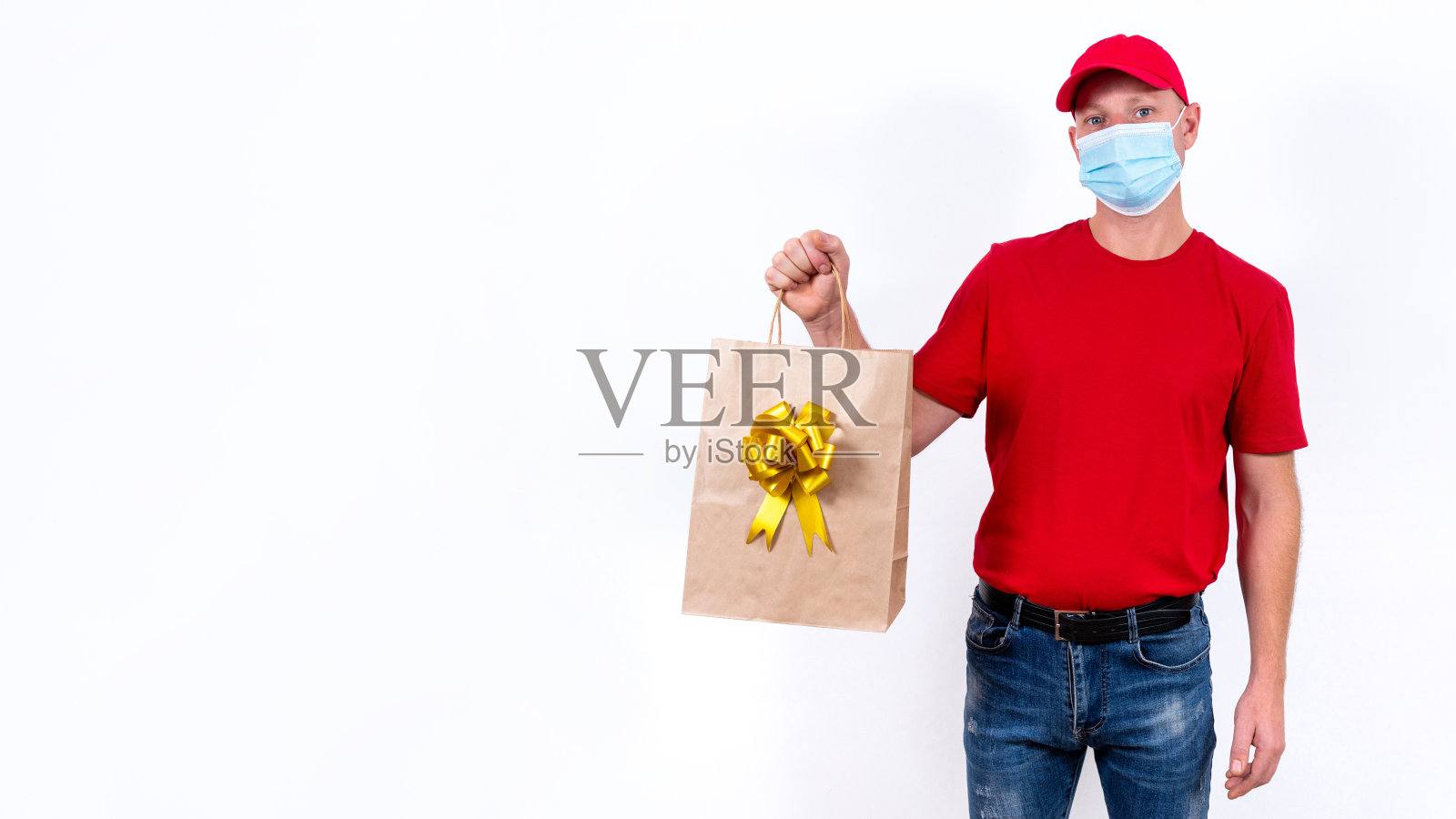 横幅。在冠状病毒大流行期间，安全非接触远程传递节日礼物。一名身穿红色制服、戴着医用防护口罩的快递员手持一个带蝴蝶结的漂亮礼品纸袋。副本的空间。照片摄影图片