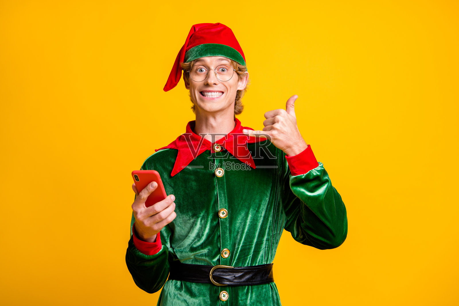 照片的家伙拿着智能手机手显示手机露齿的微笑戴着精灵天鹅绒绿色服装头饰眼镜孤立的黄色背景照片摄影图片