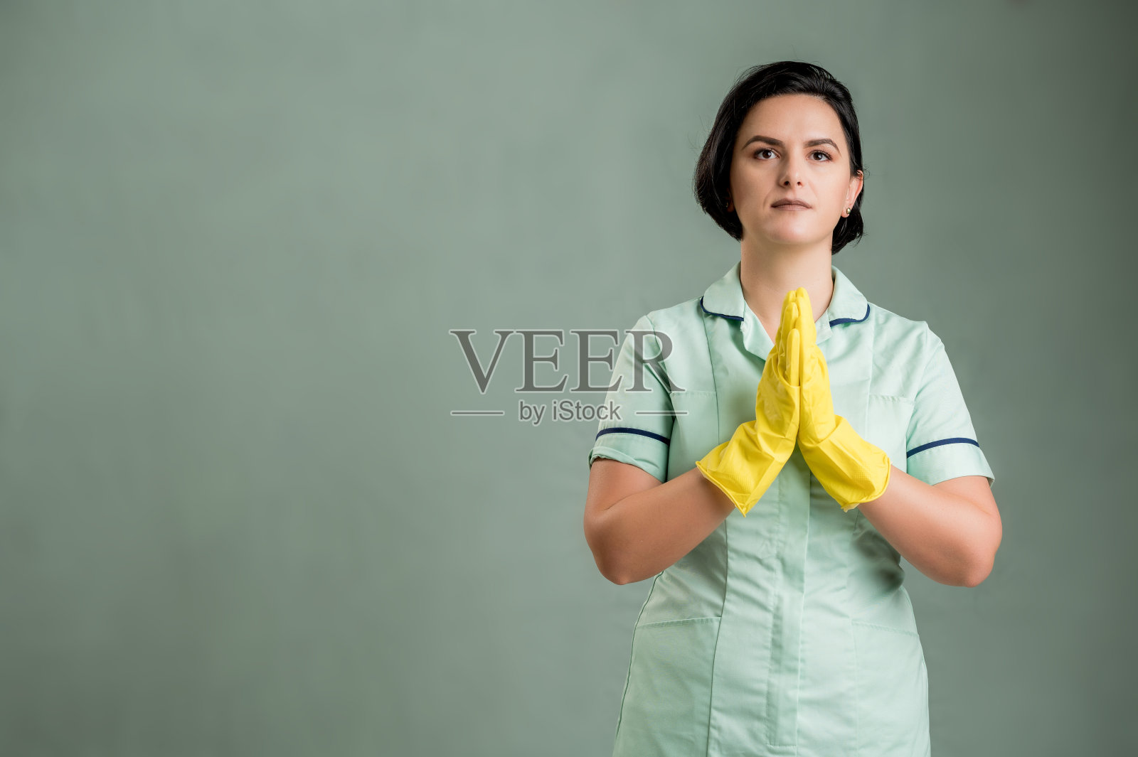 年轻的清洁女工穿着绿色的衬衫和黄色的手套祈祷照片摄影图片