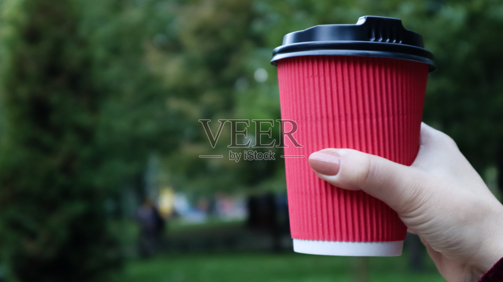 一个穿着外套的年轻女孩在公园里散步时手里拿着早上的咖啡。手握纸杯咖啡在绿色公园。结论卡布奇诺。特写,复制空间照片摄影图片