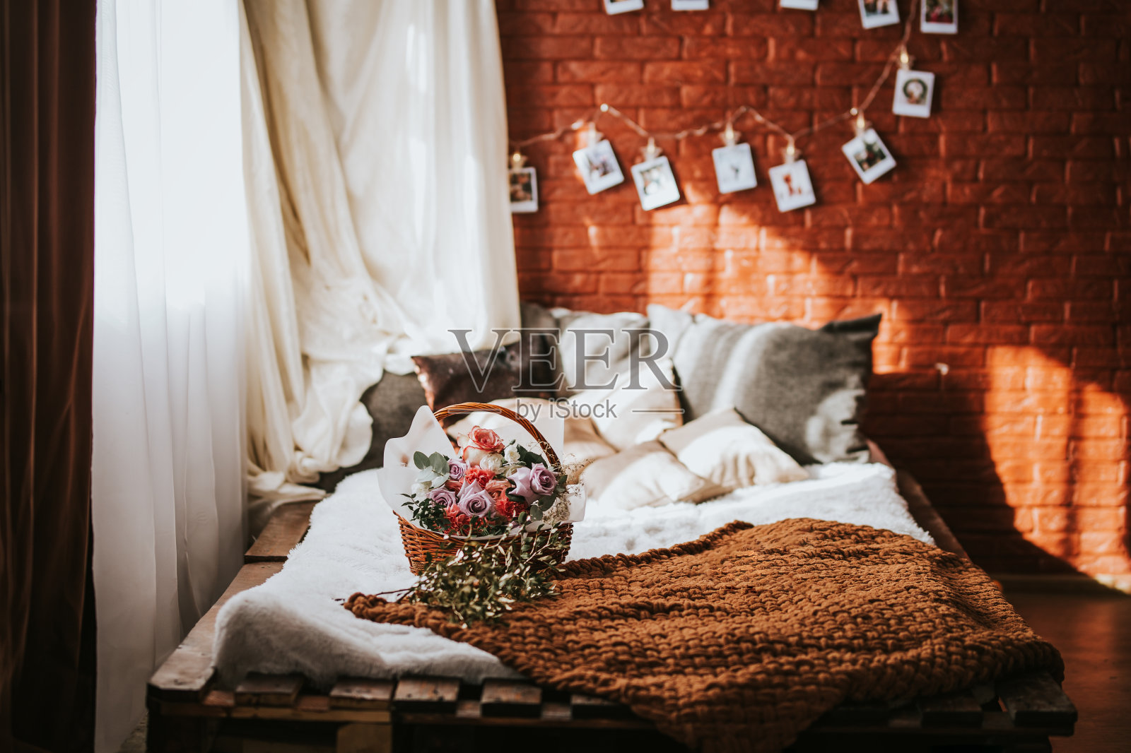 一张大床的托盘和枕头，在床上有一个大篮子的不同类型的花，一个花环的照片在砖墙上，阳光透过窗户选择焦点照片摄影图片