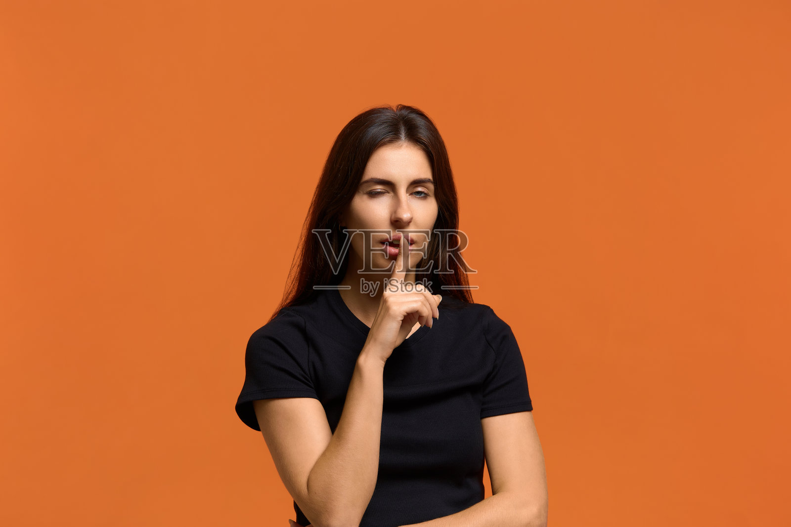 嘘，到我身边来。和穿着黑色t恤的漂亮白种女人调情，把食指放在嘴唇上，眨眼。微笑着表示沉默。孤立在橙色背景上。照片摄影图片