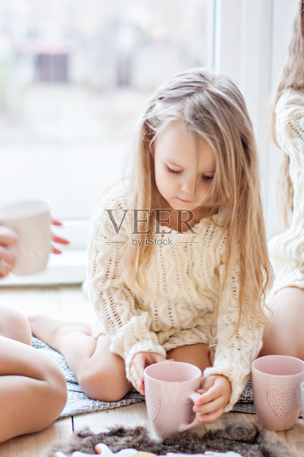 一个穿着白色针织毛衣的小女孩喝着热巧克力，可可。婴儿望着窗外，等待一个新年奇迹圣诞老人，吃着圣诞姜饼和棉花糖人。卡照片摄影图片