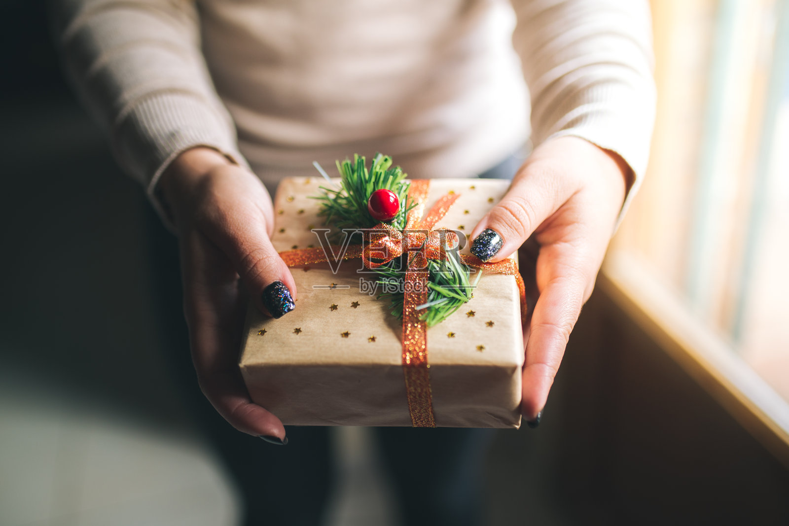 年轻的女人用闪亮的指甲拿着圣诞礼物盒并在镜头前展示它。手握新年礼盒。用牛皮纸、松枝、红丝带、星星装饰。家人礼物的概念。照片摄影图片