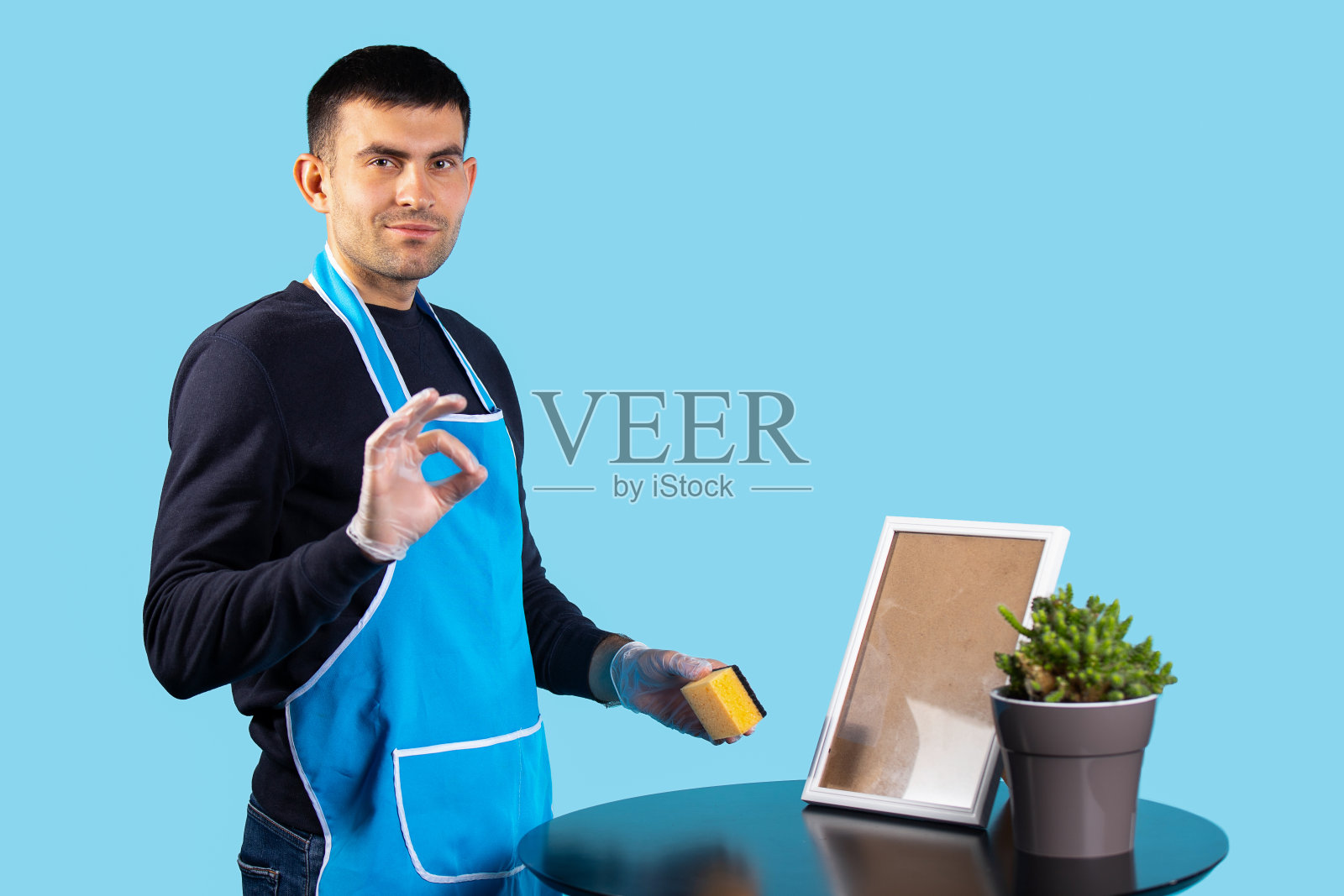 一位穿着围裙、戴着手套的英俊男子，擦去他的热情，心满意足地看着镜头。照片在蓝色的ok标志上。性别刻板印象的概念照片摄影图片