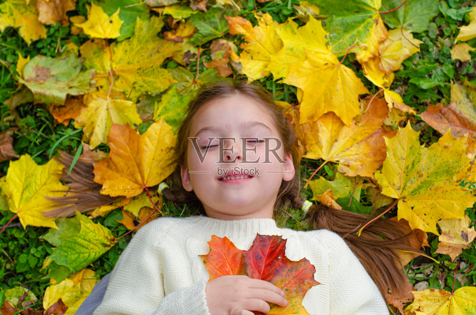 一个穿着白色针织连衣裙的小女孩微笑着躺在落叶上。秋天公园里明亮的黄色、橙色的枫叶。照片摄影图片
