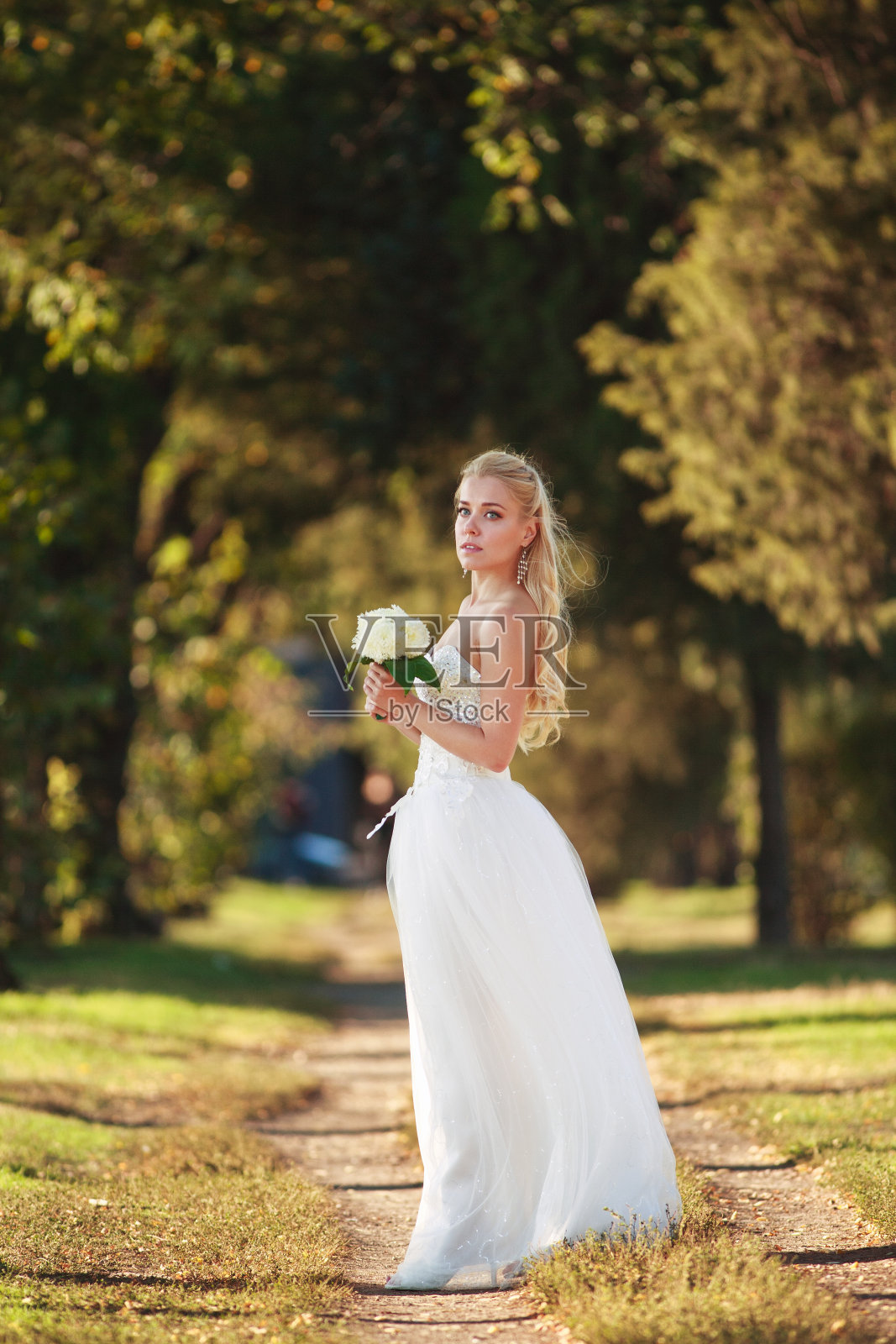 身穿白色婚纱的年轻新娘手持花束在森林公园里照片摄影图片