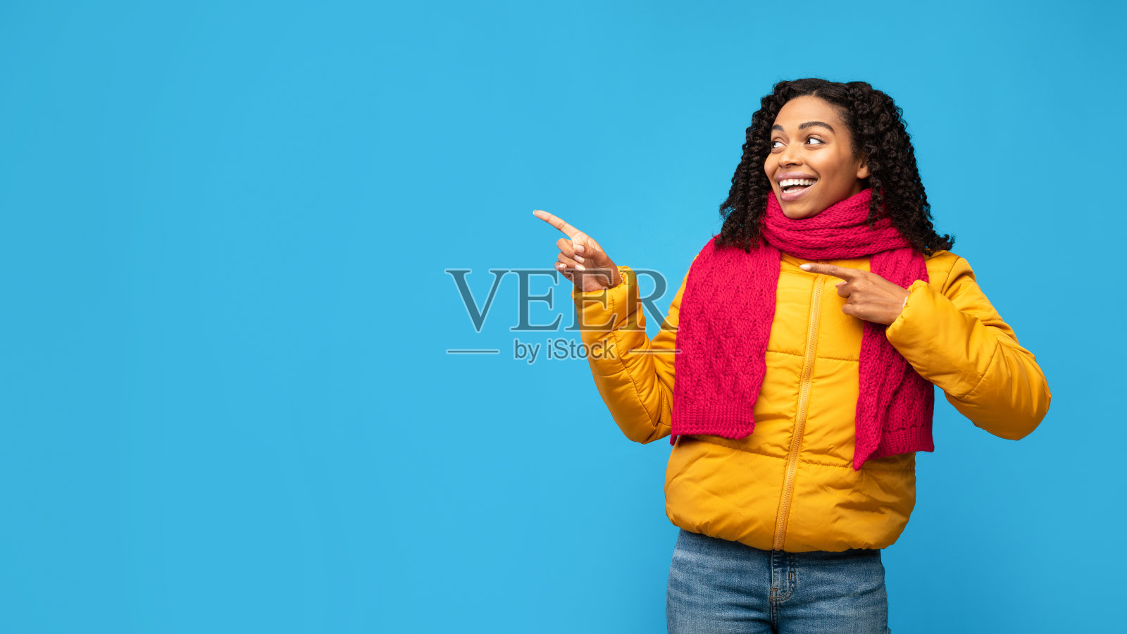 非洲女士在冬天的衣服指向一边的手指，蓝色的背景照片摄影图片