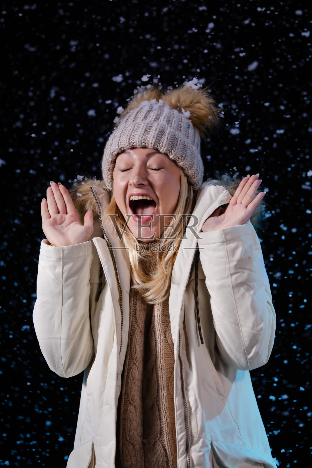 一个年轻女子在享受降雪的肖像。在黑色的背景上，一个穿着暖冬衣服的金发女郎为第一场雪而高兴。近距离照片摄影图片