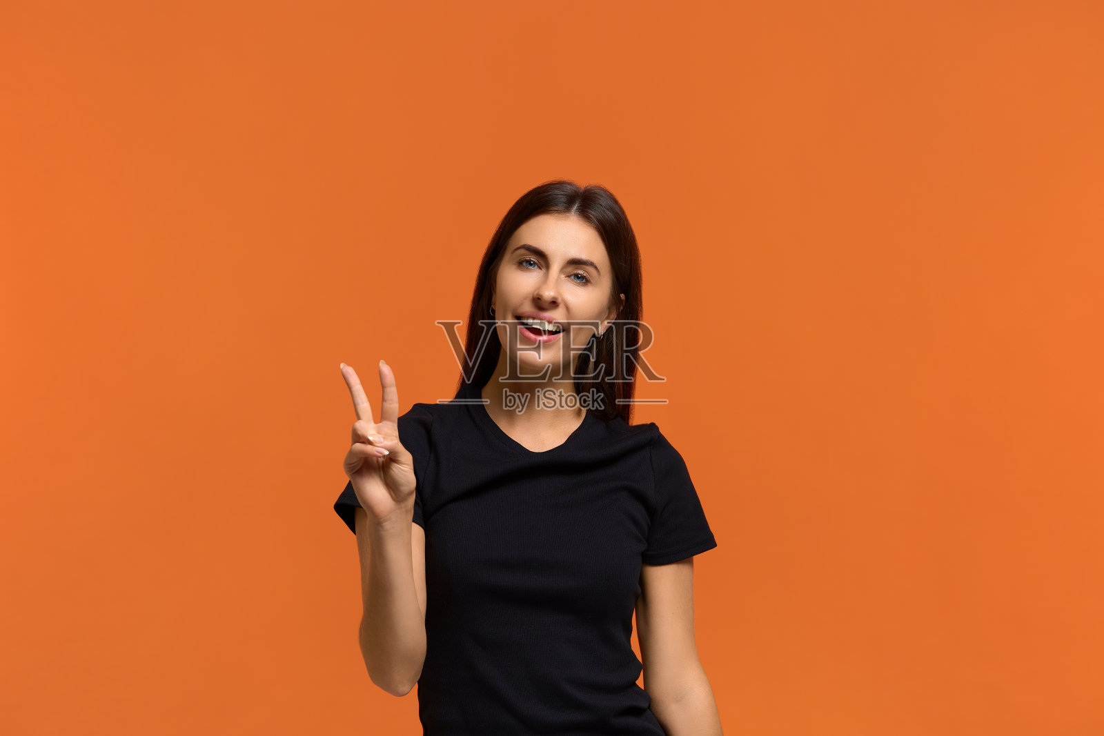 胜利的手。美丽的白人妇女在黑色t恤微笑，给和平的标志，高兴看到有人。广泛的微笑。孤立在橙色背景上照片摄影图片