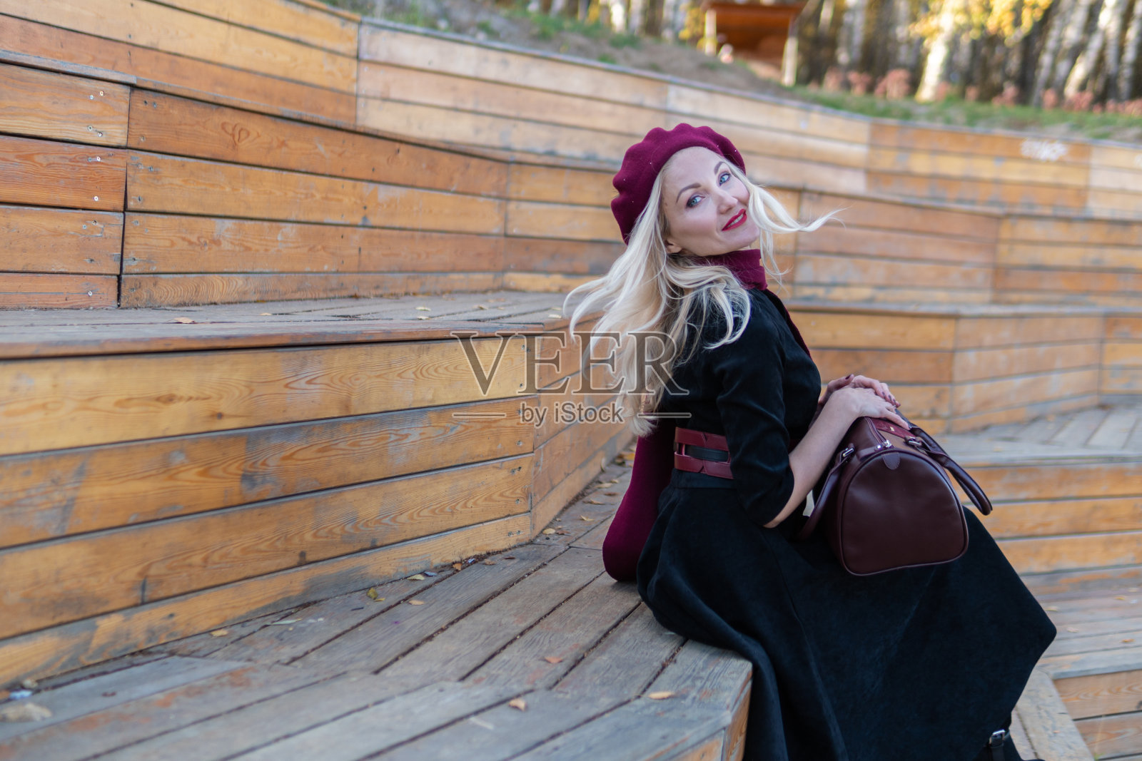 一个微笑的女孩坐在夏季剧院的木凳上，她穿着紫红色的外套和短上衣，一个成年人在蓝天的映衬下看着镜头。照片摄影图片