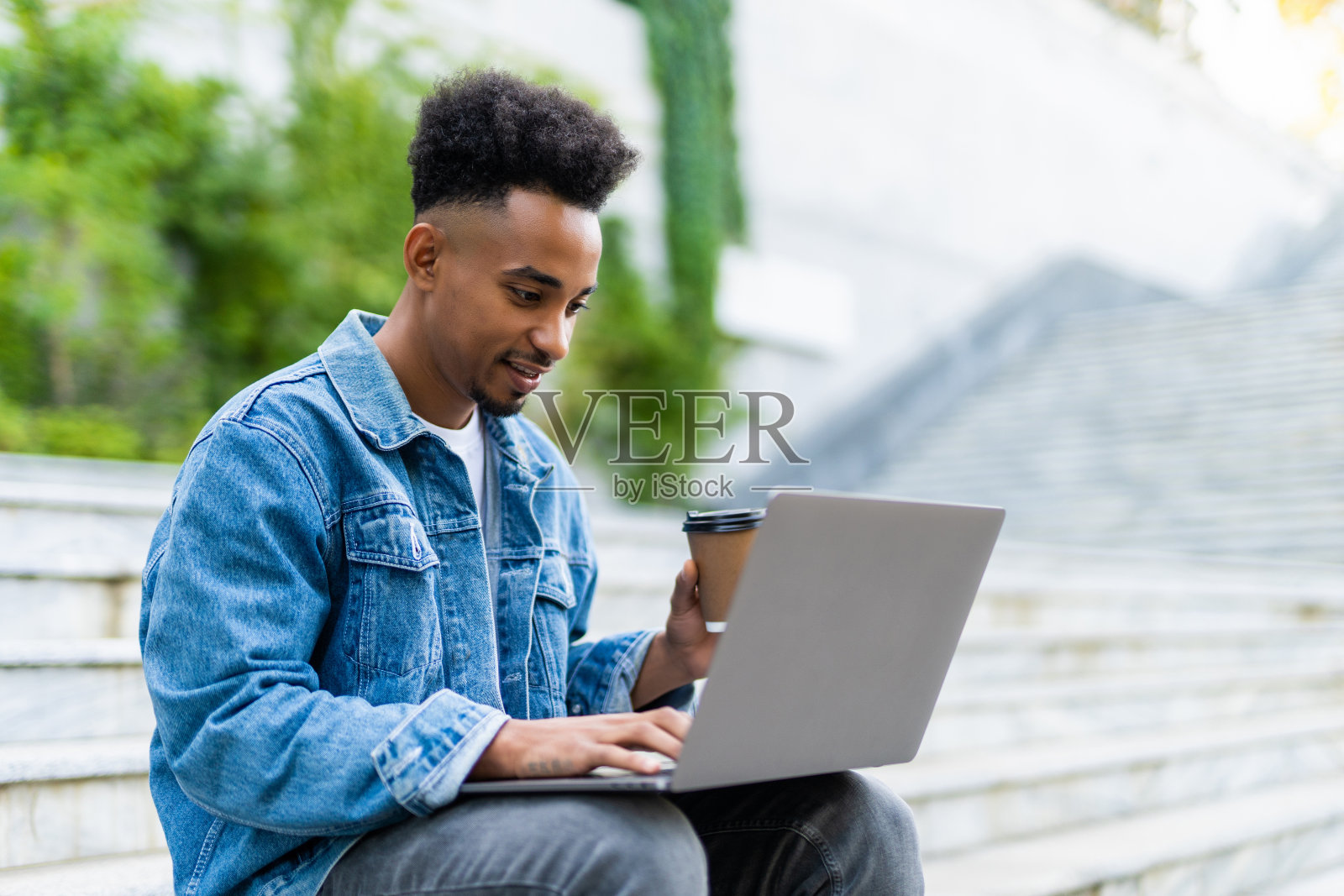 这是一幅年轻的非洲男子穿着牛仔裤夹克在户外用笔记本电脑工作的肖像，咖啡杯放在长凳上照片摄影图片