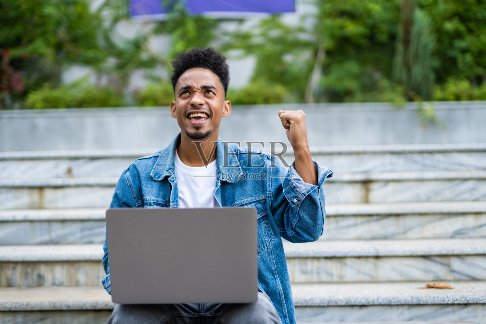 幸福成功的非裔美国人双手高举在空中。年轻随意的学生拿着笔记本电脑在户外庆祝胜利，坐在公园的长椅上。照片摄影图片