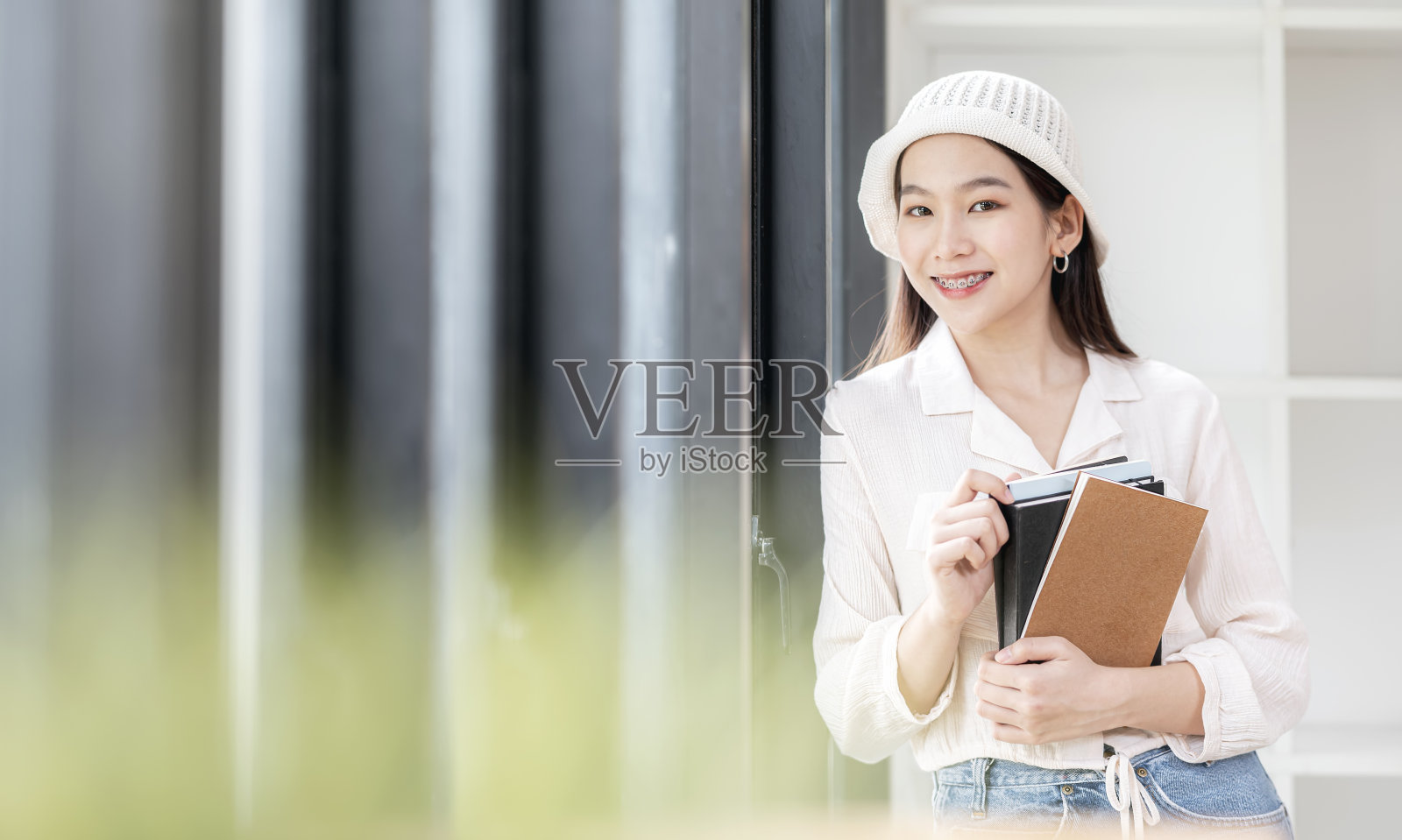 年轻女学生戴着羊毛帽，戴着牙套，拿着书站在图书馆里。照片摄影图片