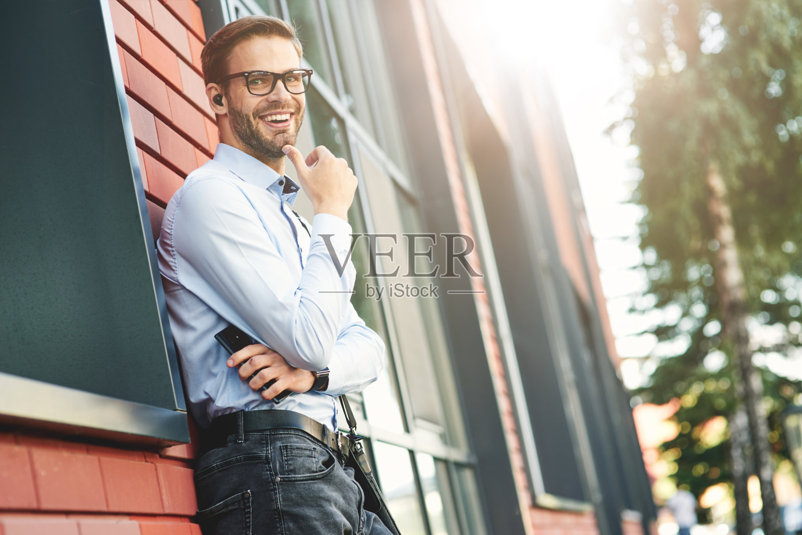 年轻快乐的商人戴着无线耳机，拿着智能手机，看着摄像头，微笑着站在户外照片摄影图片