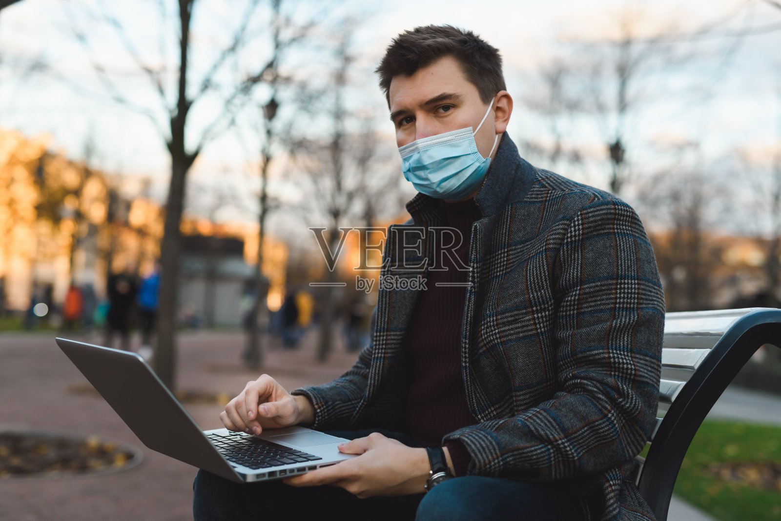 一个年轻英俊的男人，穿着秋日外套，戴着防护面罩，在户外的街道上用笔记本电脑工作。新冠肺炎大流行期间的社交距离。新冠肺炎疫情在欧洲大城市蔓延。照片摄影图片
