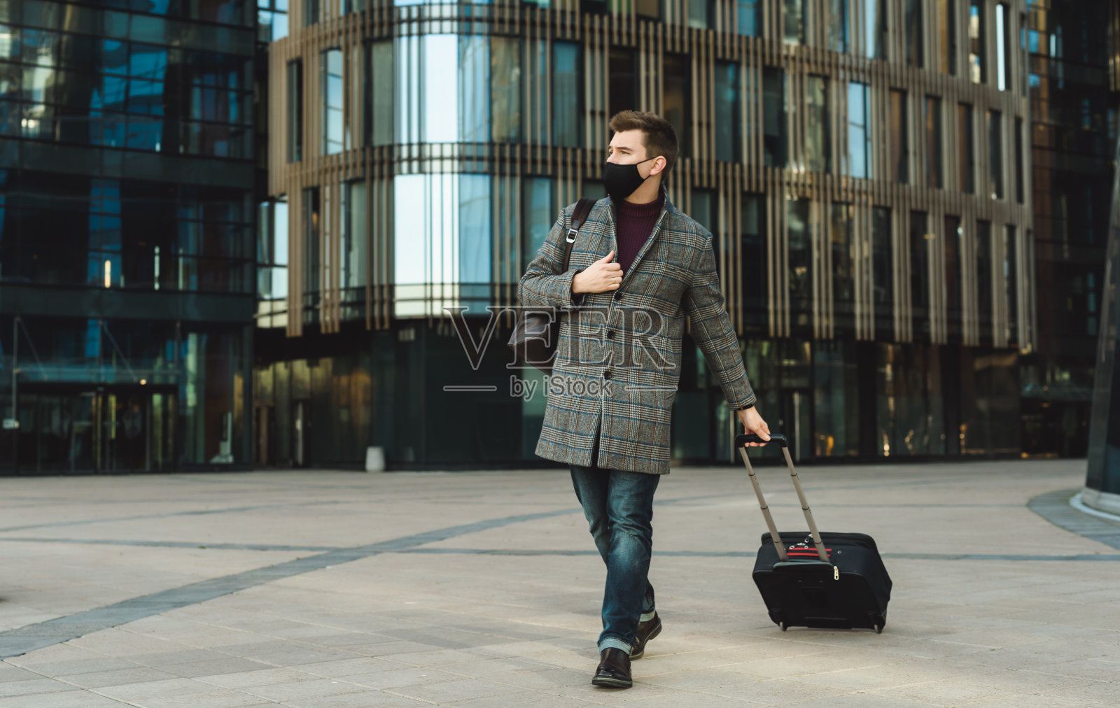 新冠肺炎疫情期间出差。年轻男子拿着行李箱背对着机场或火车站大楼。在城市街道公共场所佩戴口罩并保持社交距离照片摄影图片