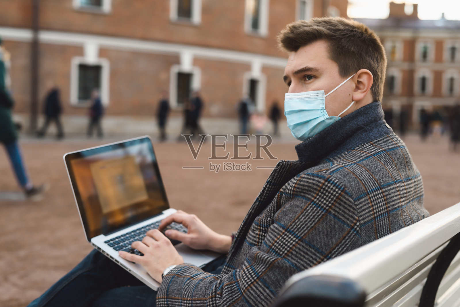 一个年轻英俊的男人，穿着秋日外套，戴着防护面罩，在户外的街道上用笔记本电脑工作。新冠肺炎大流行期间的社交距离。新冠肺炎疫情在欧洲大城市蔓延。照片摄影图片