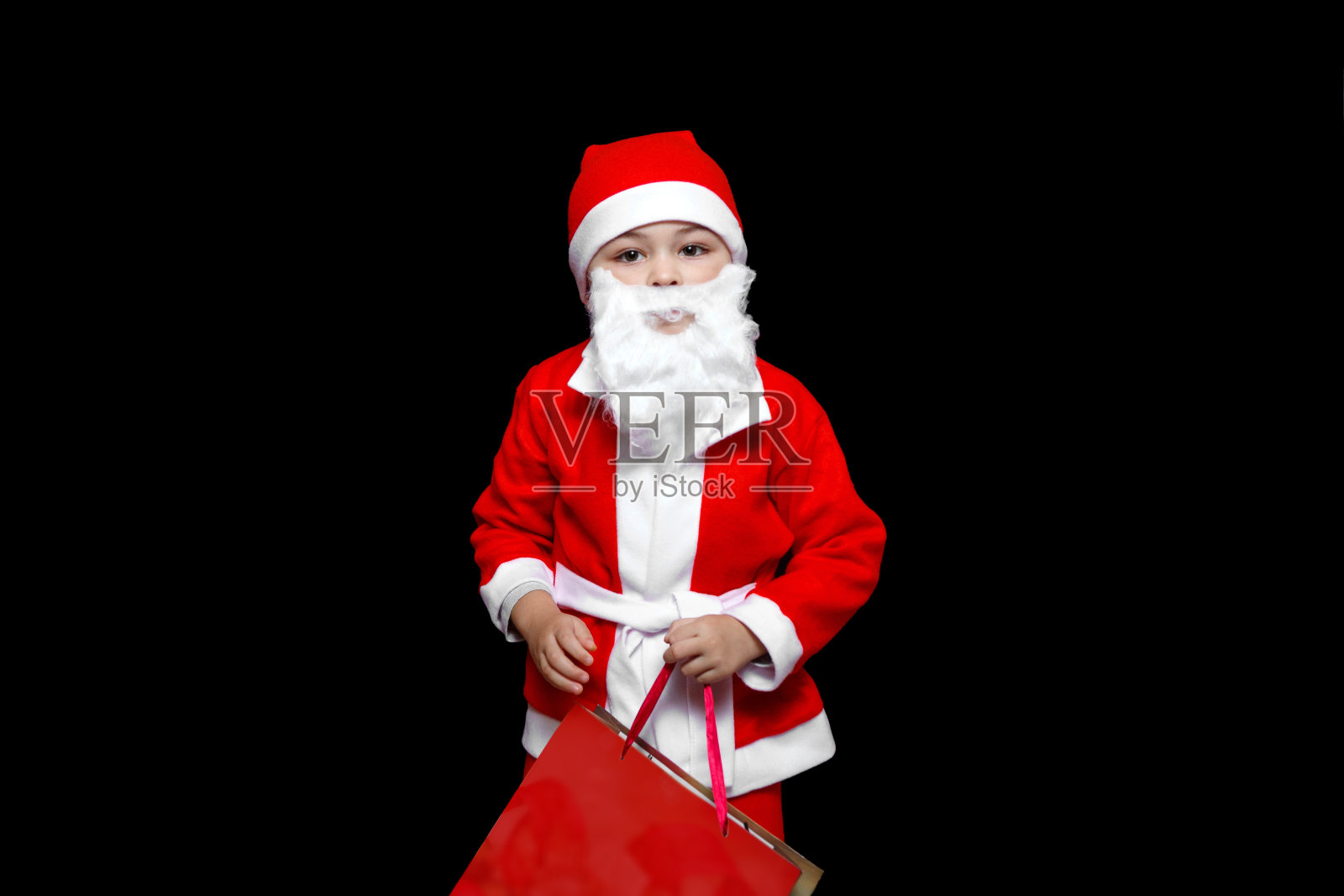 一个五岁的小男孩穿着滑稽的圣诞老人服装，拿着一大包礼物站在那里。在一个孤立的黑色背景上照片摄影图片