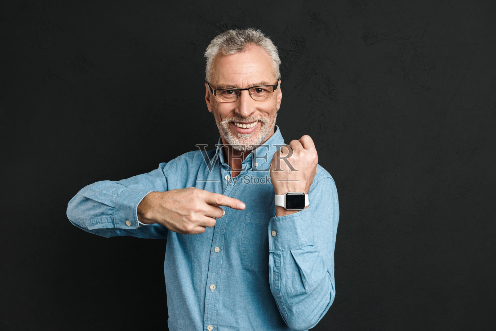 60年代的照片，留着灰白头发的绅士戴着眼镜，微笑着指着他的手表，孤立在黑色的背景照片摄影图片
