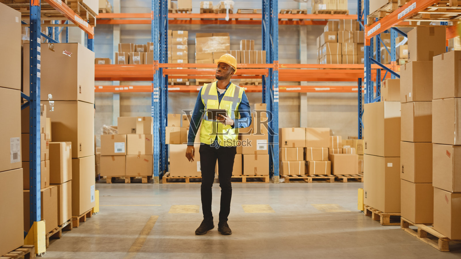 帅气的男员工戴着安全帽，手持数字平板电脑，走过摆满货架的零售仓库。在物流配送中心工作。照片摄影图片