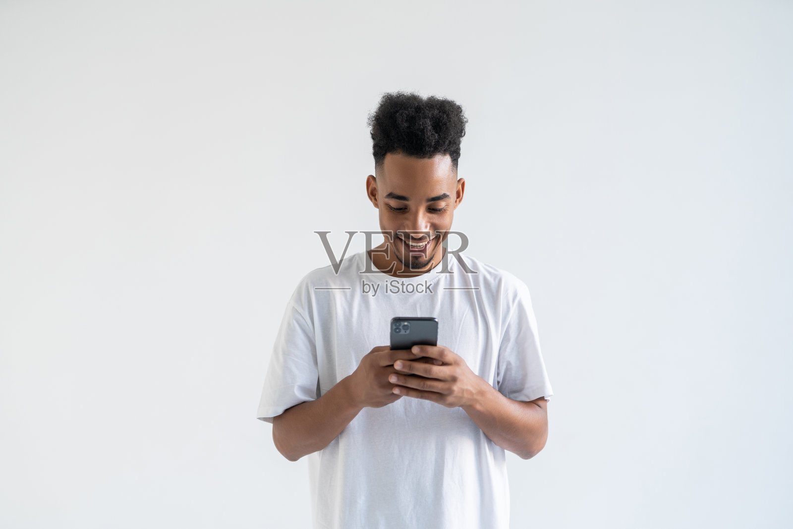 一名年轻的非洲裔美国男子身穿白色t恤站在镜头前，专注地微笑着看着他手持的智能手机屏幕照片摄影图片