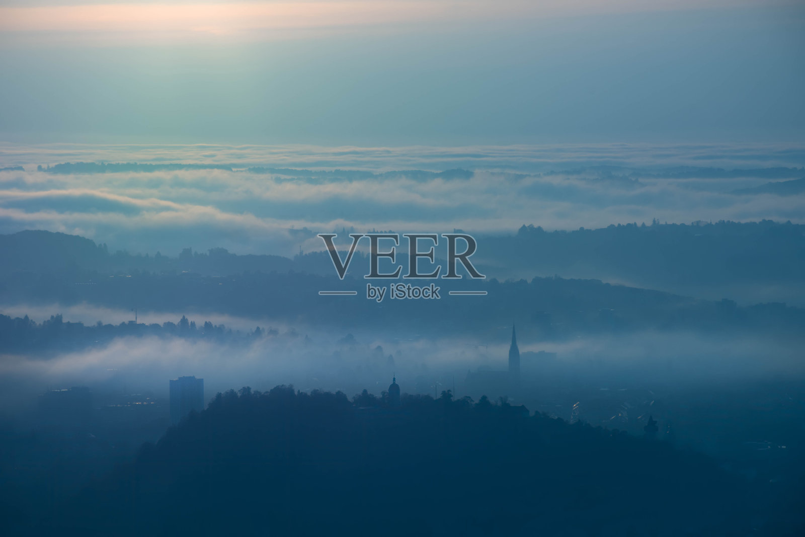 奥地利斯蒂里亚地区，格拉茨市上空雾蒙蒙的日出，有施洛斯贝格山和耶稣圣心教堂塔。秋天的早晨从Plabutsch山的全景。照片摄影图片