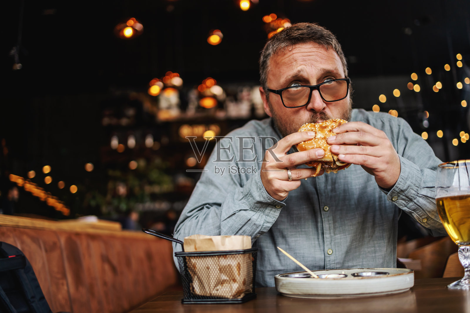 蓄着胡须的中年饥饿男子坐在餐馆里吃着美味的汉堡。照片摄影图片