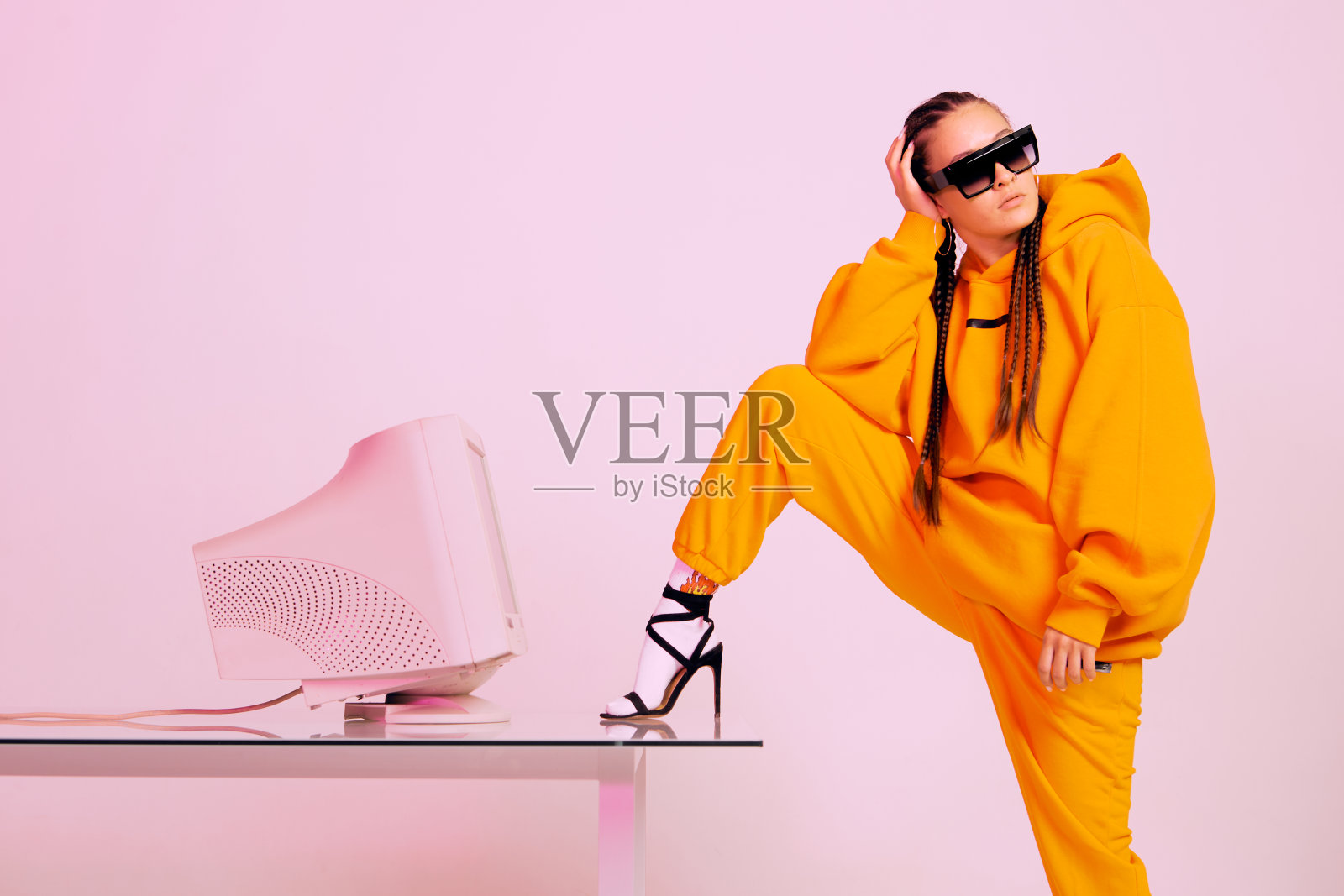 时髦的年轻女性办公室职员戴着太阳镜，戴着旧电脑显示器，脚踏在办公桌上。彩色的光照片摄影图片