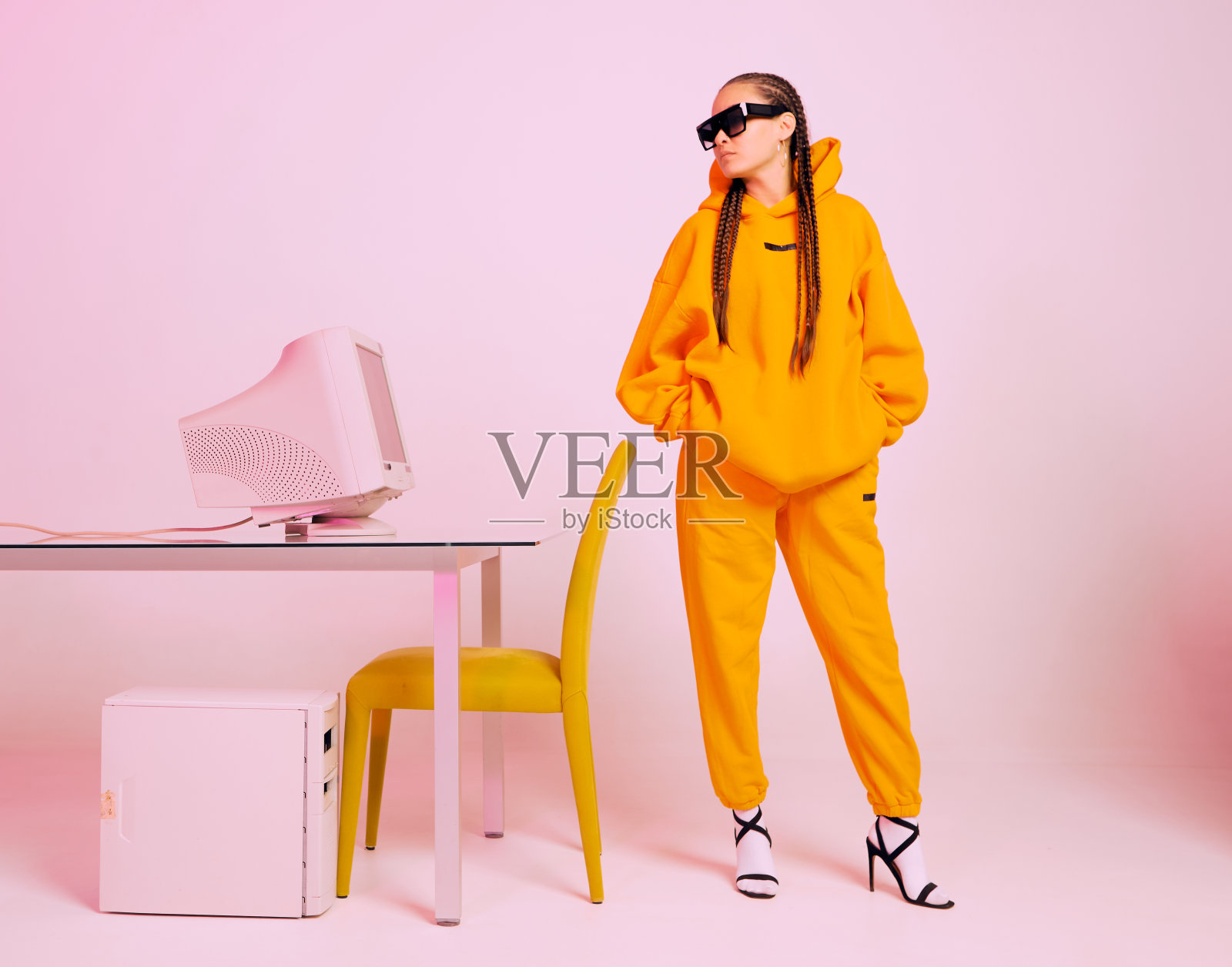 时髦的女办公室职员戴着太阳镜和帽衫站在过时的电脑前。彩色的霓虹灯照片摄影图片