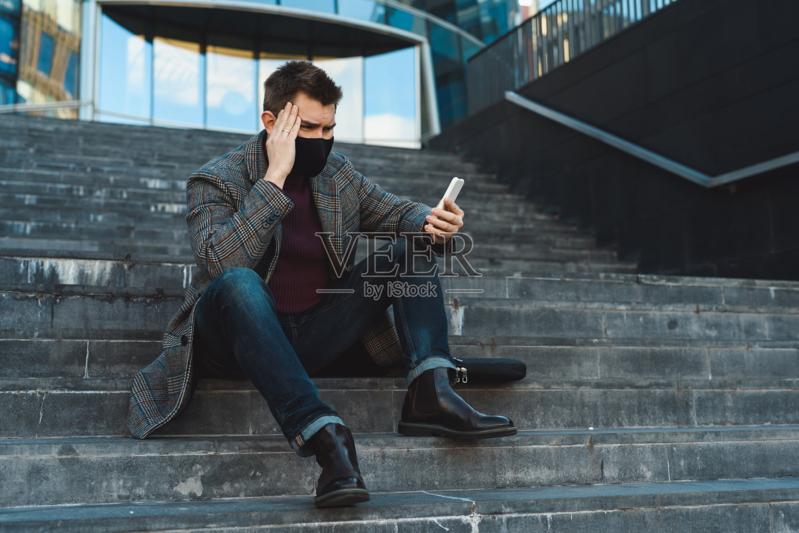 一名年轻的上班族在新冠肺炎疫情期间失业。一个年轻的商人拿着智能手机受苦。大城市里的社交距离。在公共场所戴口罩照片摄影图片