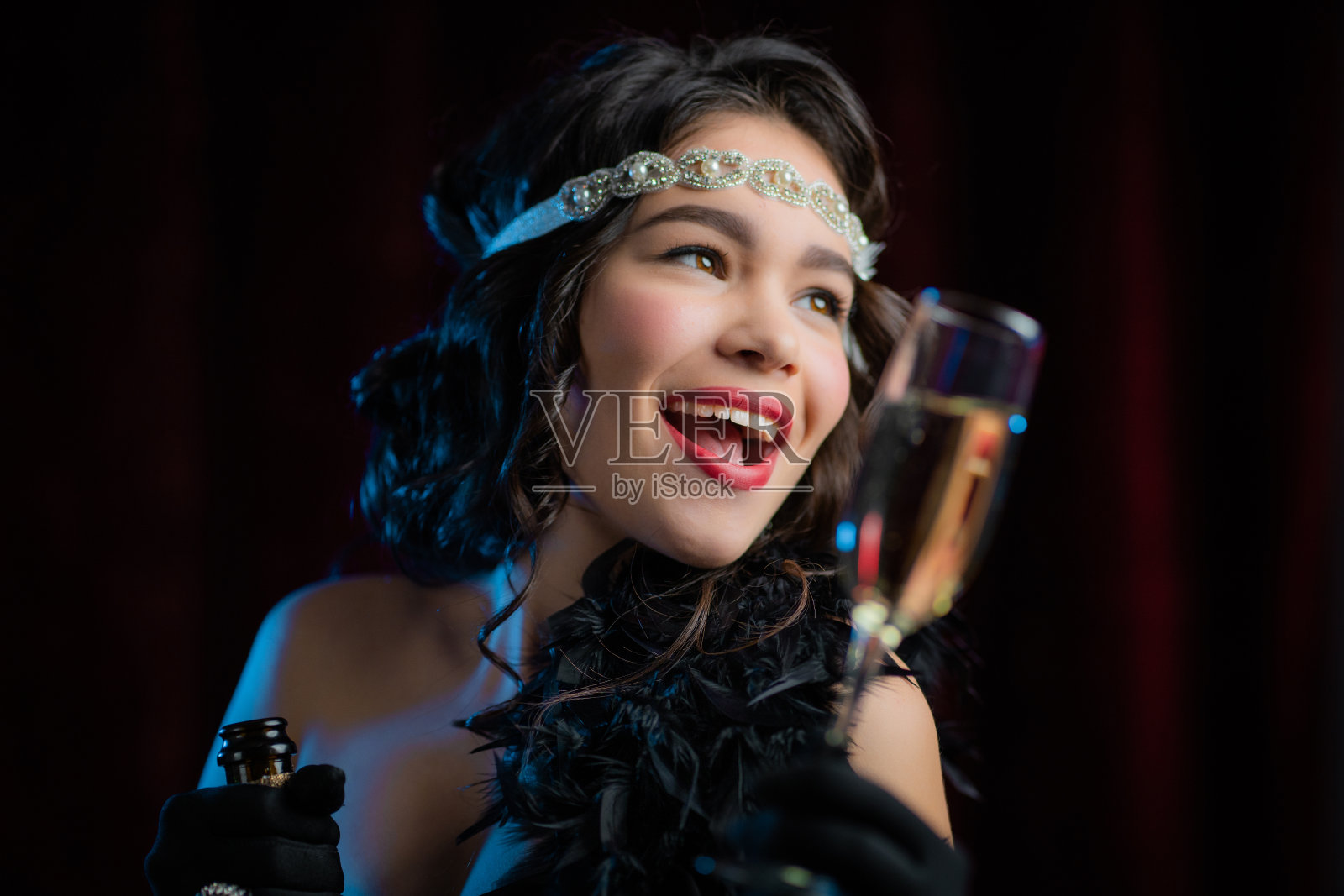 复古风格的女人穿着Flappers的连衣裙举起一杯香槟像吐司，喝葡萄酒在天鹅绒的背景。喧嚣的二十年代，复古，派对，时尚概念照片摄影图片