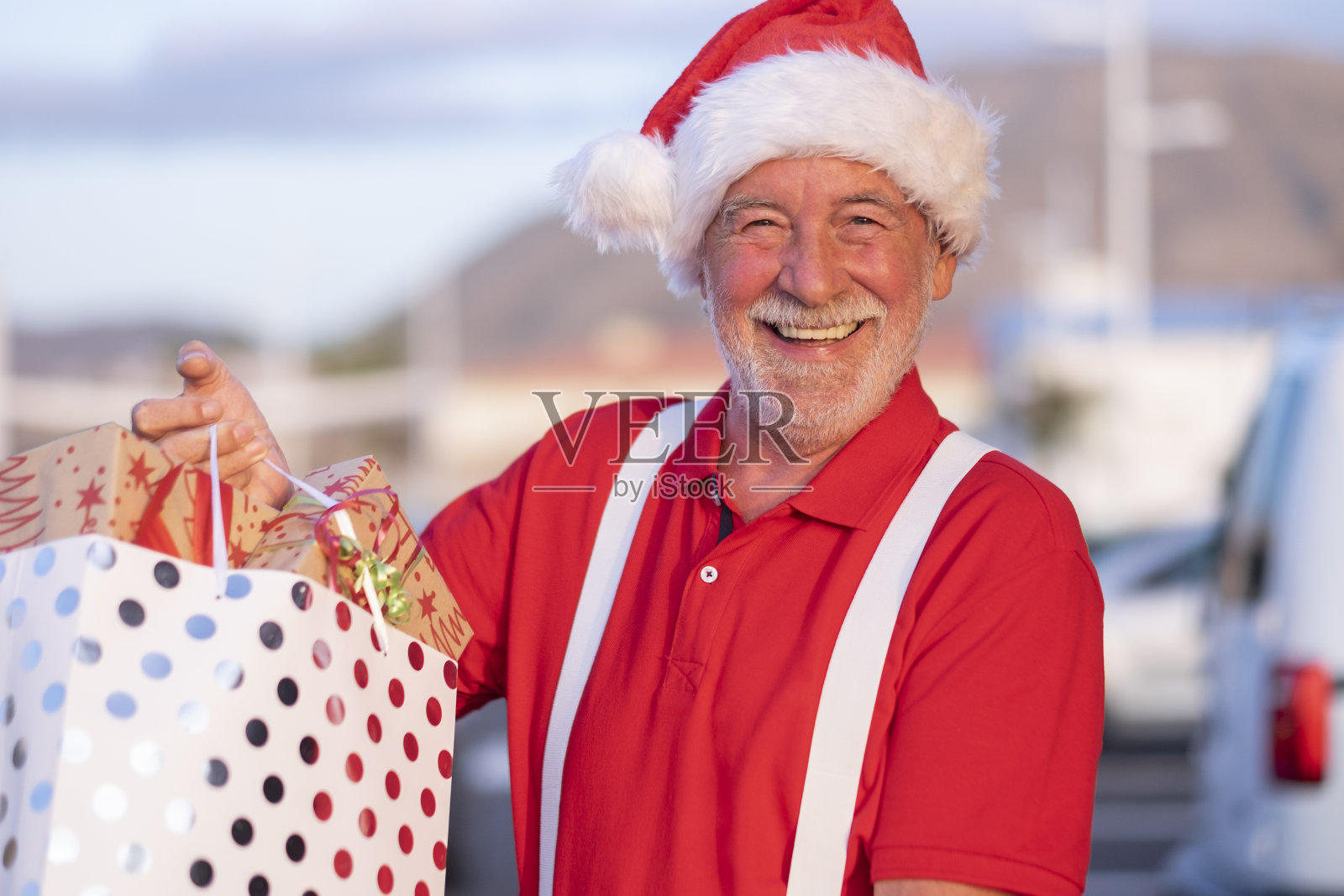 一个微笑的大胡子老人的肖像购物圣诞老人的帽子和吊带，拿着一个购物袋满礼物和看着相机。快乐的退休人员照片摄影图片