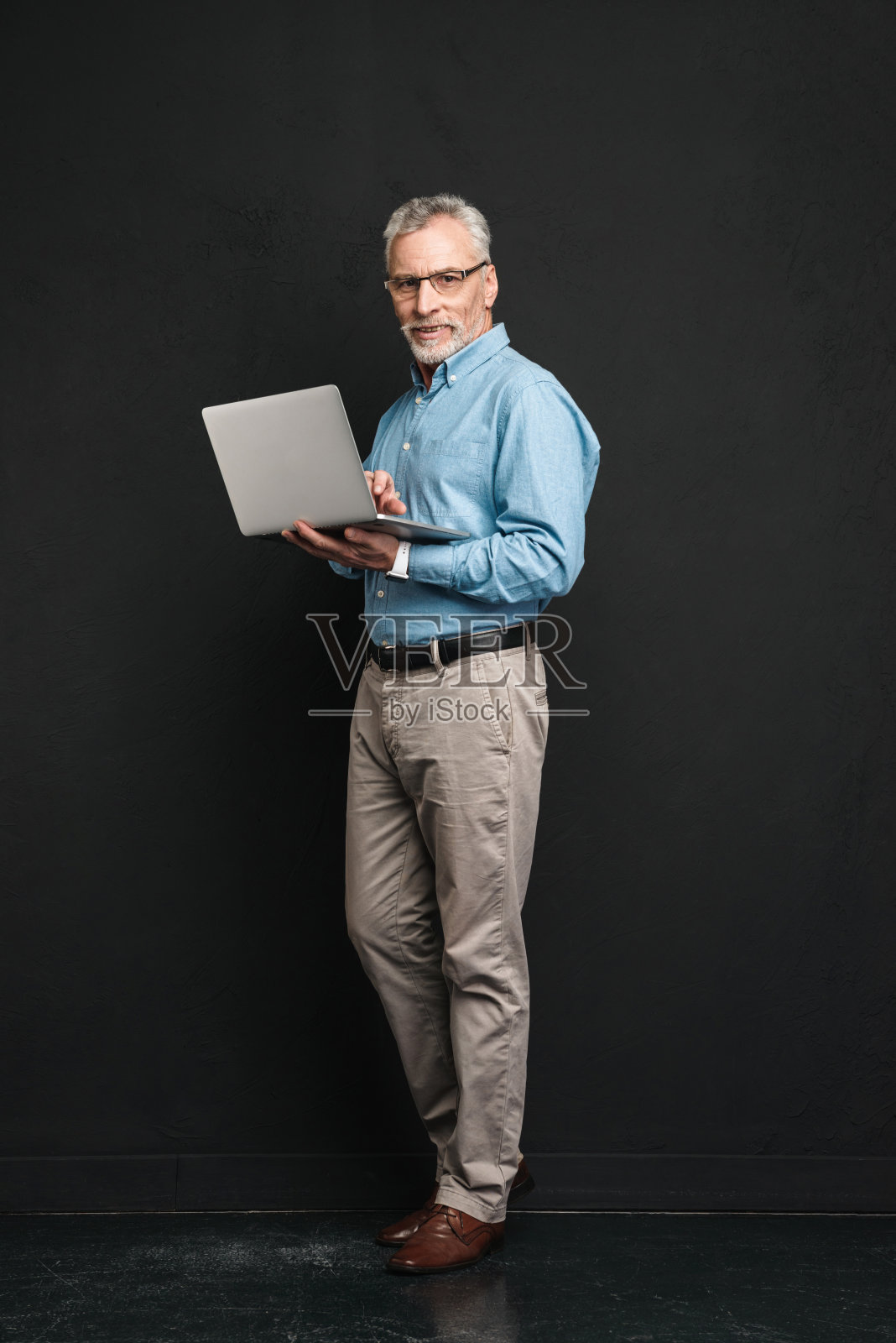 全长照片上的老商人70年代与灰色的头发和胡子工作，而手里拿着银色的笔记本电脑，孤立在黑色背景照片摄影图片