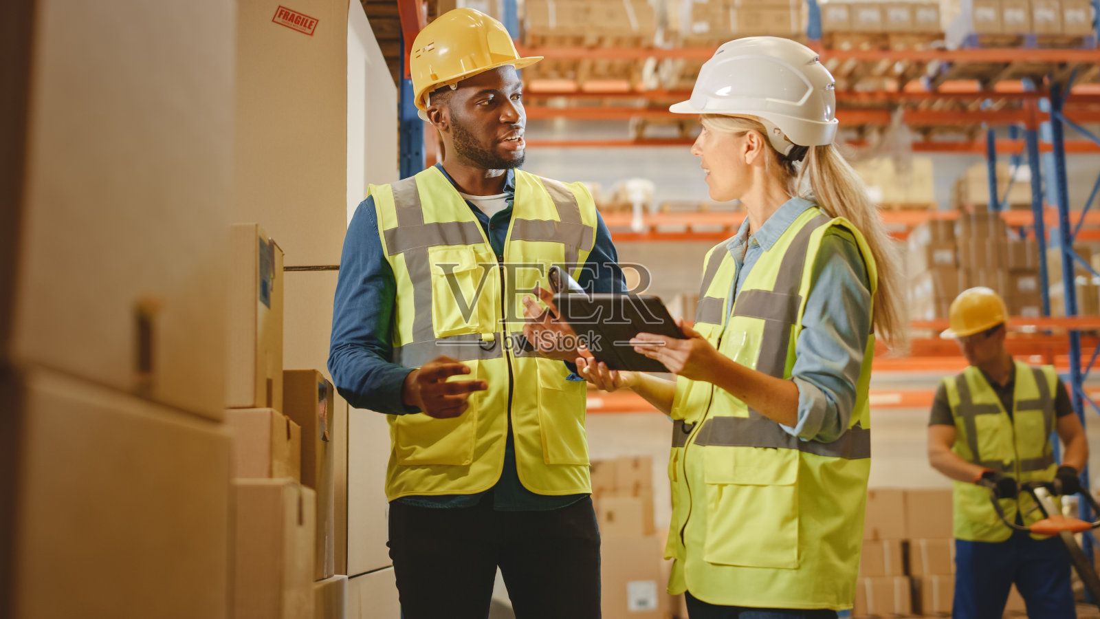 零售仓库满货架的货物在纸板箱，男工人和女主管持有数字平板电脑讨论产品交付，同时扫描包裹。销售物流中心照片摄影图片