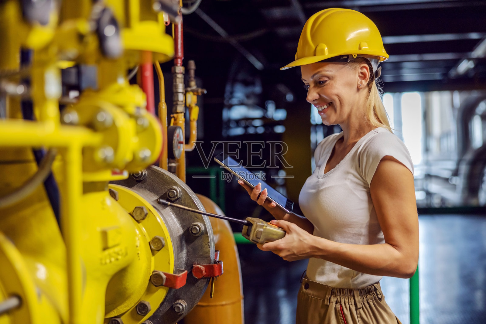 女主管，穿着工作服，头戴头盔，面带微笑，工作勤奋，站在加热车间，手拿平板，检查管道上的热量。女性做男性工作的概念。照片摄影图片