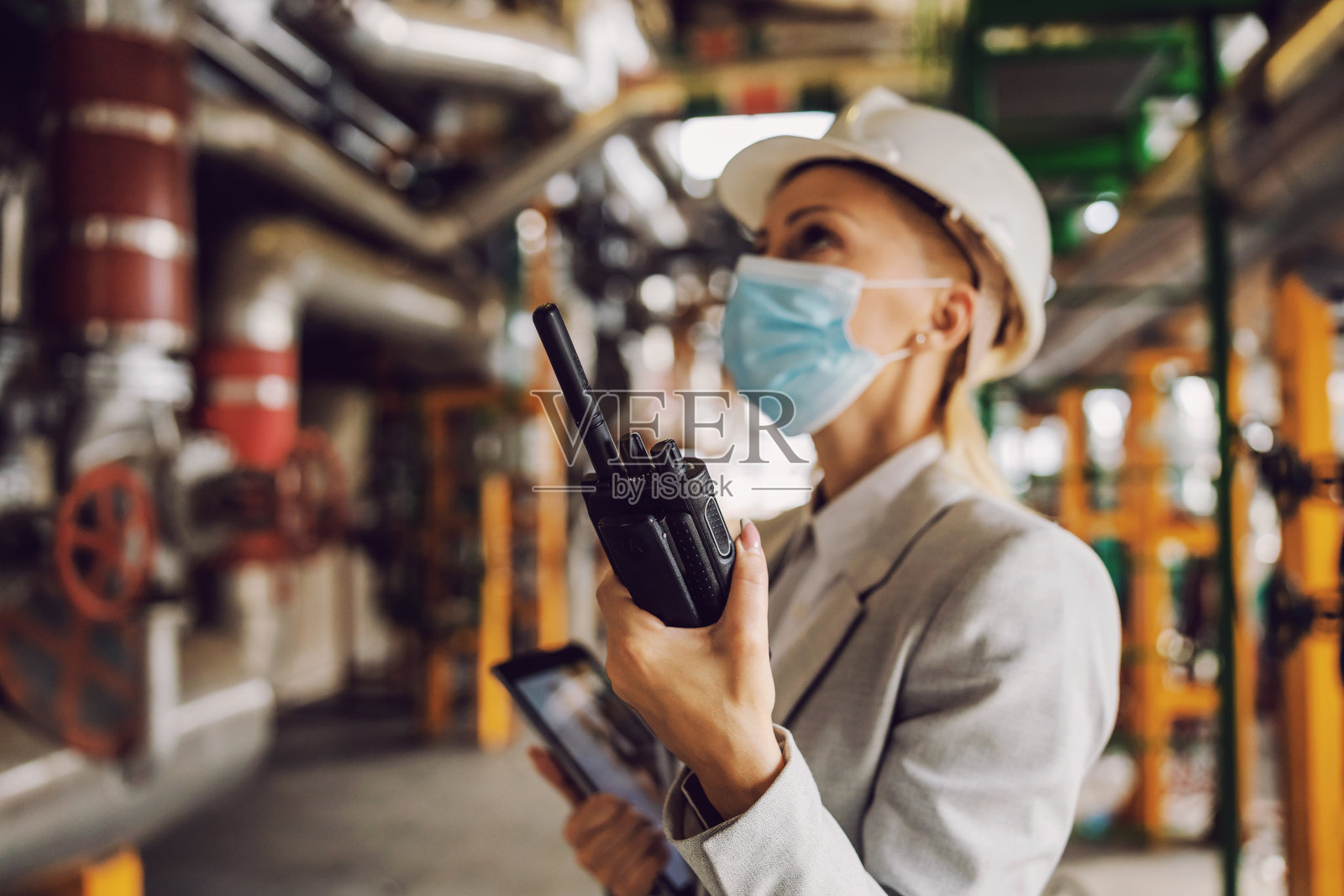 中年、经验丰富、工作努力的女主管，穿着西装、戴面罩和头盔，手持平板电脑，用对讲机讲话，站在加热厂应对冠状病毒爆发。照片摄影图片