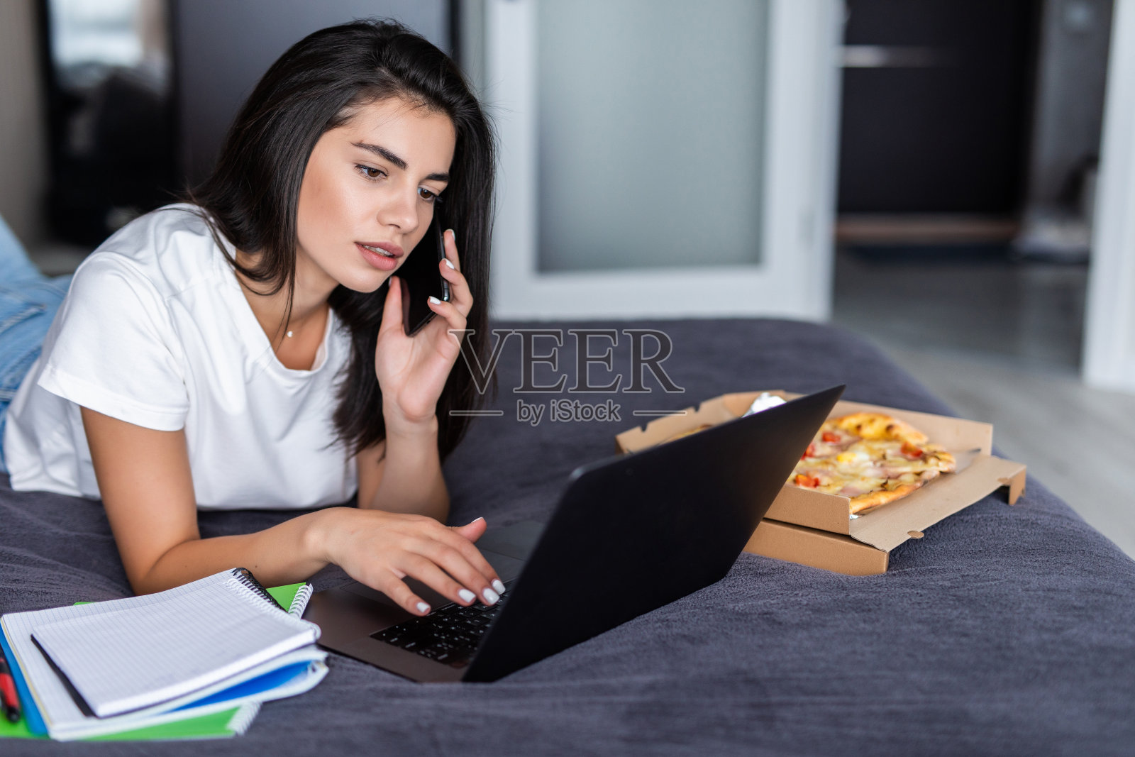 一个年轻的女人坐在床上，拿着笔记本电脑，拿着一盒披萨打电话照片摄影图片