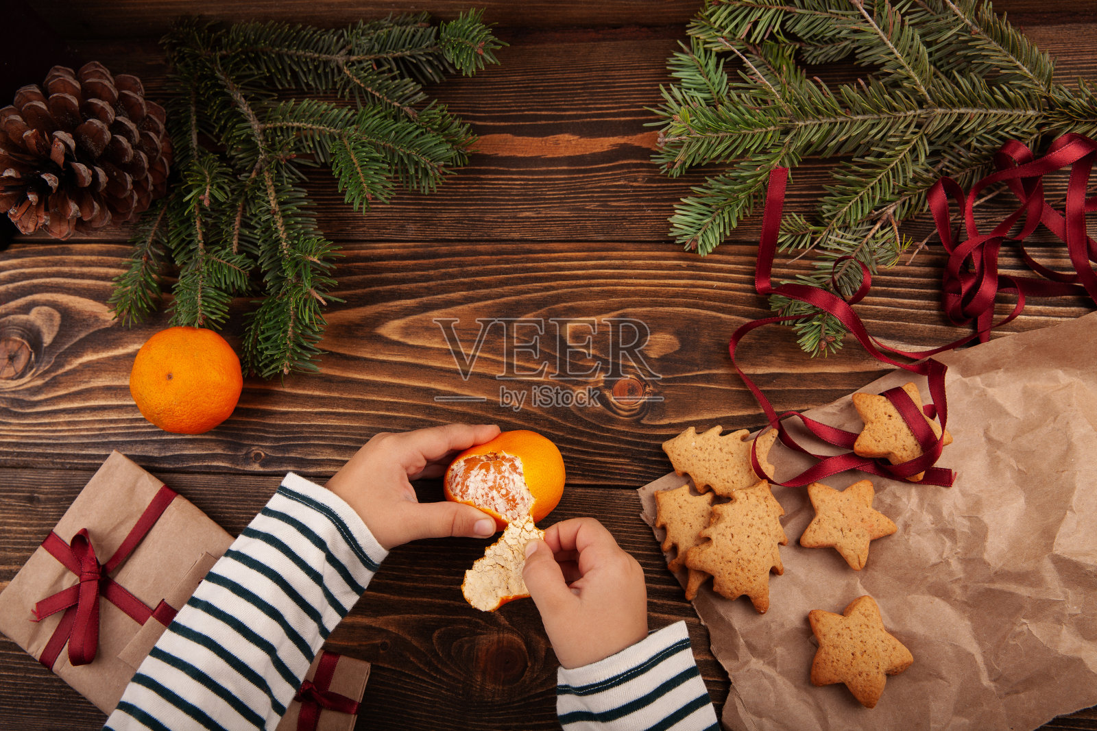 俯视图的孩子剥橘子或桔子在木桌上。桌子上有圣诞饼干和礼物。照片摄影图片