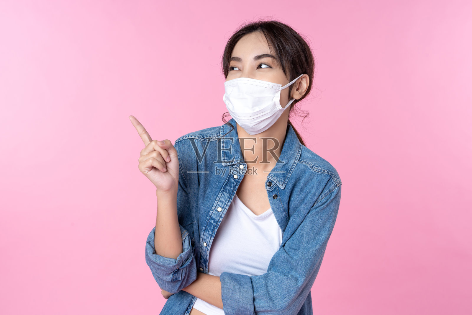 年轻快乐的亚洲女子手指向上复制空间戴口罩，以防止感染冠状病毒，她牛仔夹克和背着背包的衬衫拍摄在孤立的粉红色背景照片摄影图片