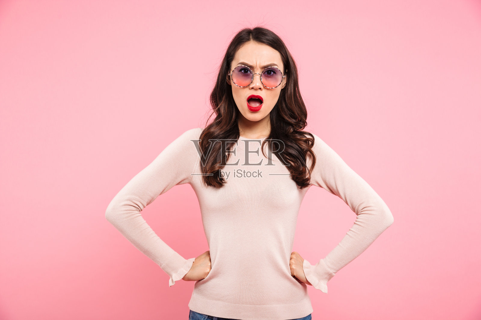 照片中愤怒的女人不经意地牵着手放在腰上，愤怒地看着镜头，在粉红色的背景下被孤立照片摄影图片