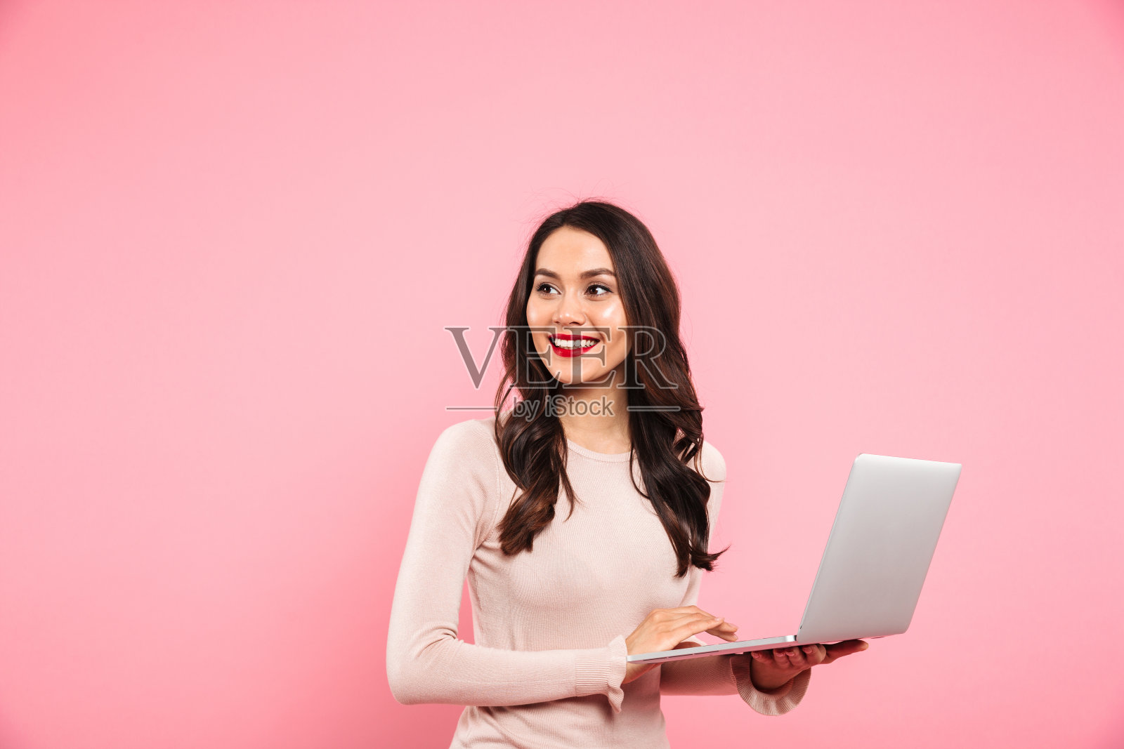 照片的黑发可爱的女人拿着银色的笔记本电脑，看着旁边的copyspace与真诚的微笑，孤立在粉红色的背景照片摄影图片