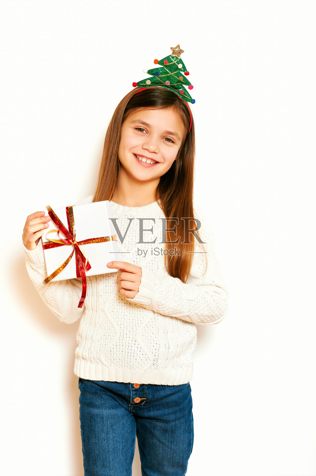 工作室的圣诞肖像年轻甜美的小女孩在白色的背景，穿着冬天的套头衫和圣诞树发带，拿着包装的礼物照片摄影图片