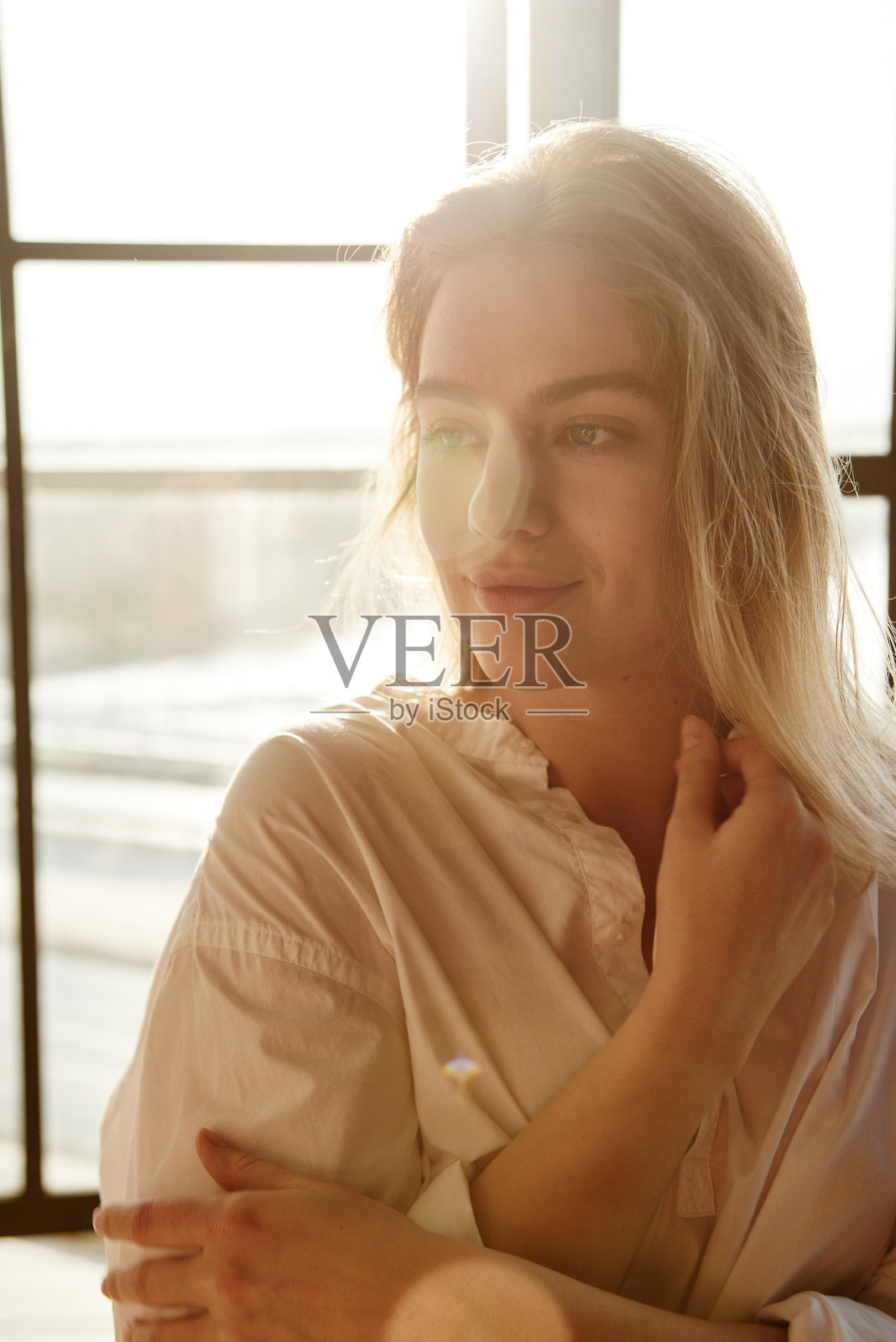 在一扇窗户的背景上，一个可爱的金发女郎穿着一件白色男子衬衫，温柔地微笑着，神秘地望着远方照片摄影图片