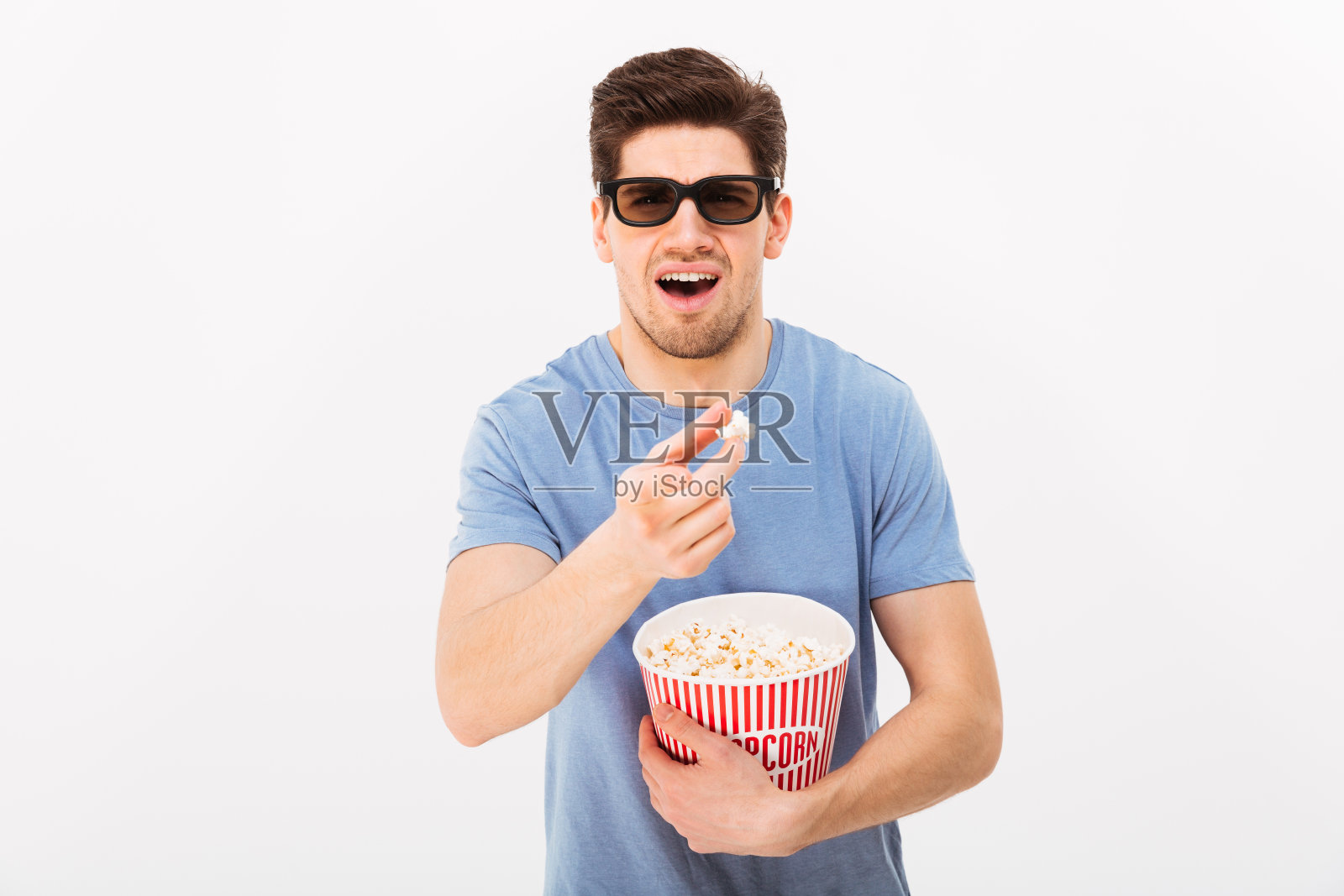 帅气的男人穿着休闲t恤，戴着3d眼镜，拿着爆米花桶，手指对着镜头，孤立在白色的墙壁上照片摄影图片