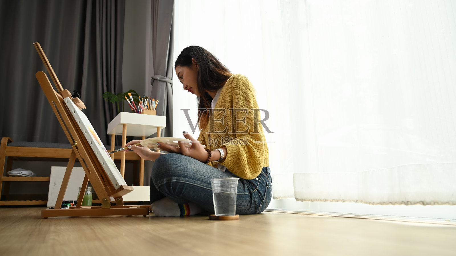 一个年轻的女画家坐在画布前的地板上，在她的工作室画画的侧视图。照片摄影图片